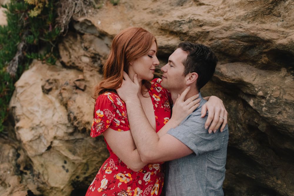 Couple in romantic embrace near rocky cliff, woman in red floral dress with auburn hair, man in gray shirt.