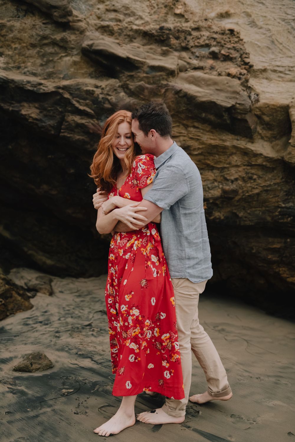 Couple embracing barefoot on a beach, woman in red floral dress, man in blue shirt, rocky cliffs behind them.