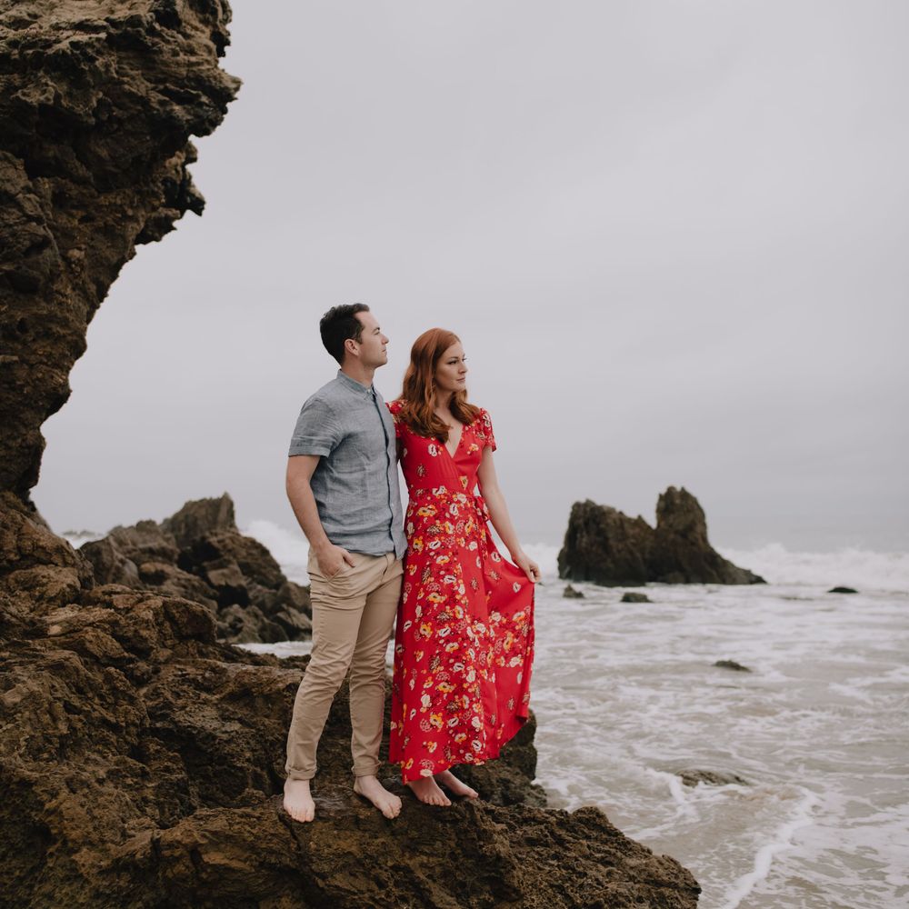Couple standing on rocky coastal cliffs, woman in red floral dress, man in gray shirt, with ocean waves in background.