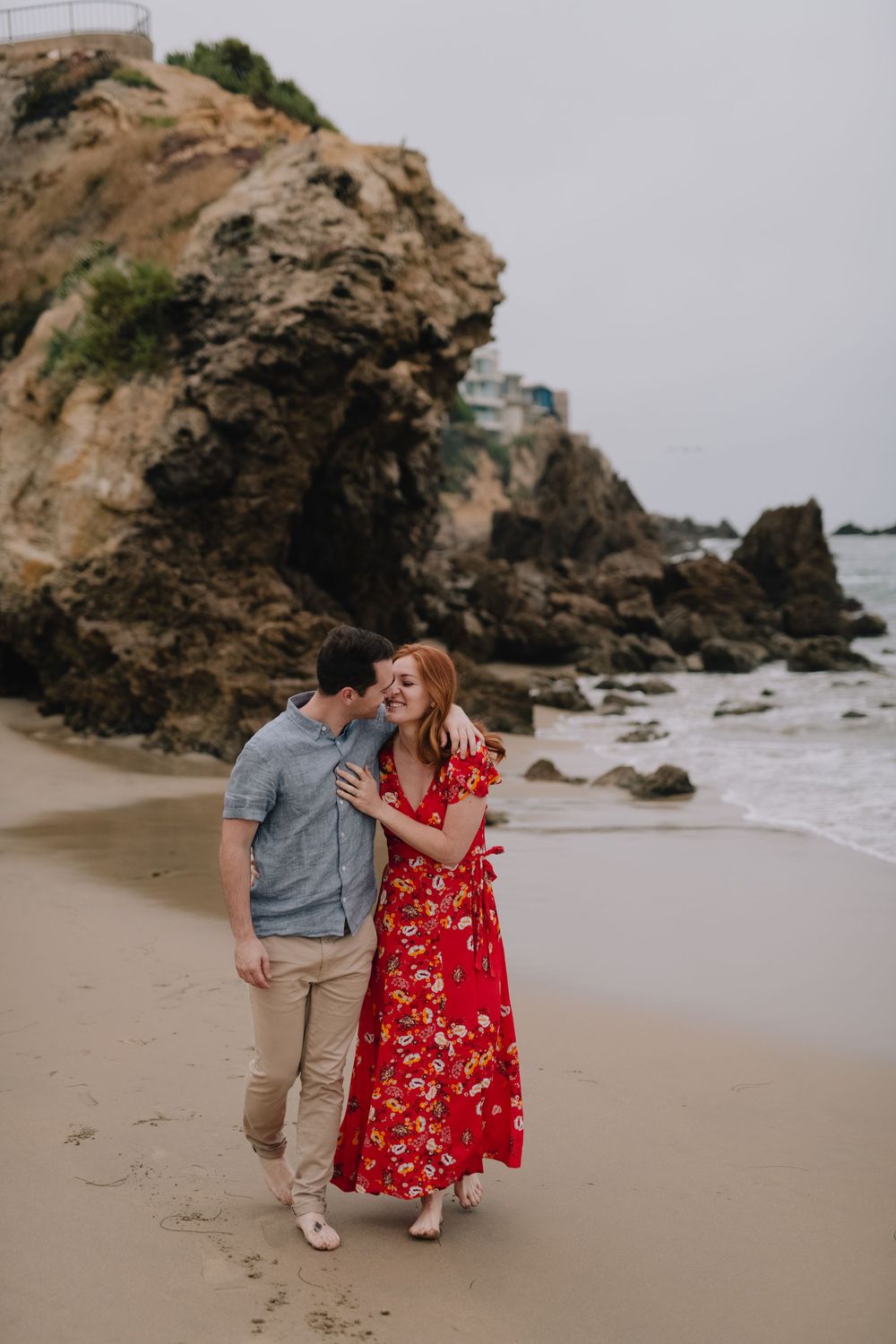Couple walking on a rocky beach, woman in red floral dress, man in blue shirt, smiling together near ocean cliffs.