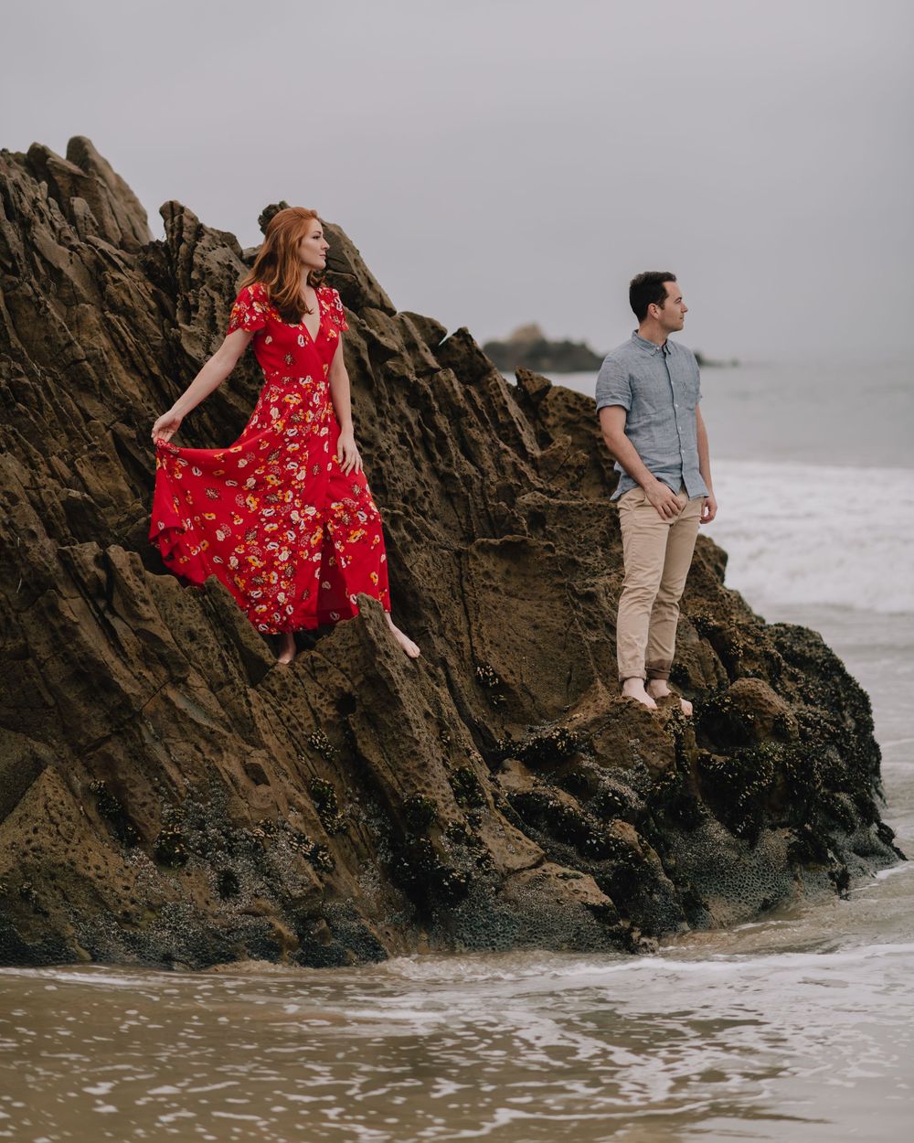 A couple poses on rocky coastal cliffs, woman in red floral dress and man in casual attire, with ocean waves below.