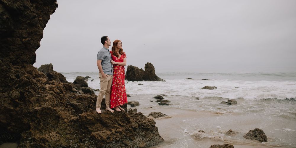 Couple embracing on rocky beach with crashing waves, woman in red floral dress, moody overcast sky backdrop.