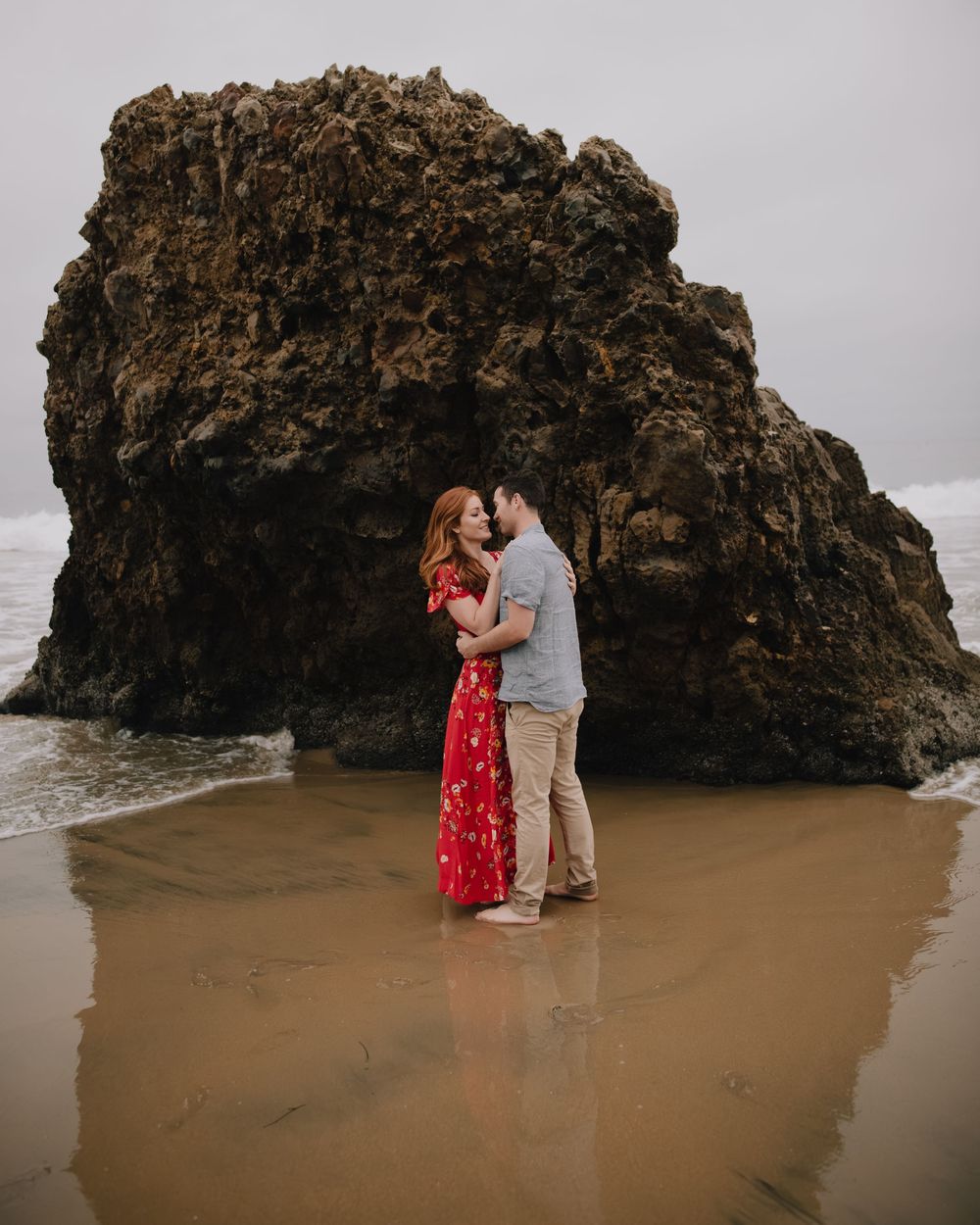Couple embracing romantically on a sandy beach with a dramatic rock formation and ocean waves behind them.