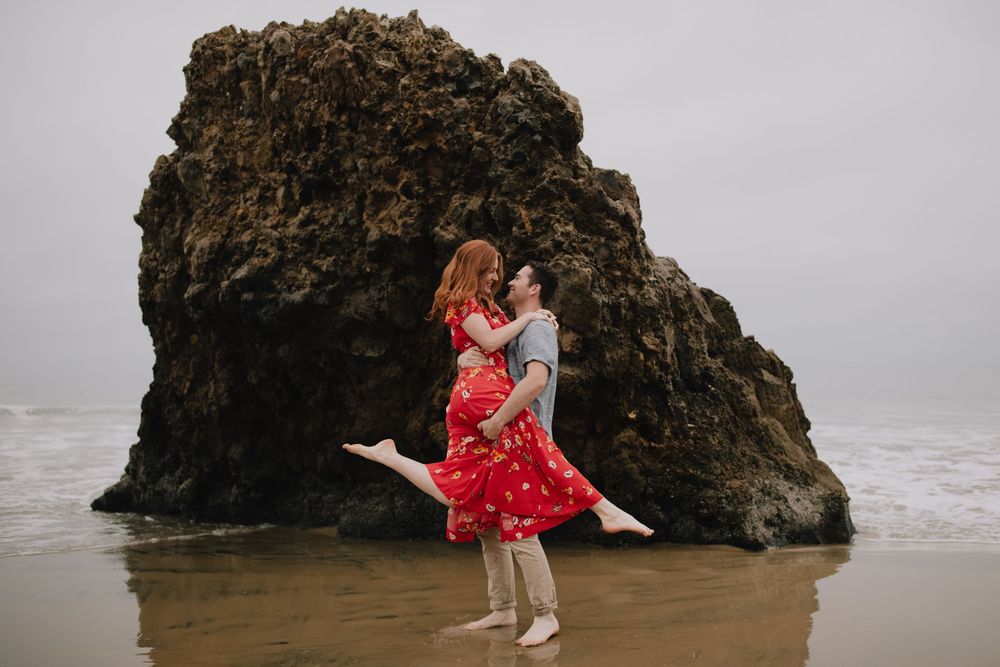 Couple embracing on a misty beach with a large rock formation, woman in red dress lifted by partner.