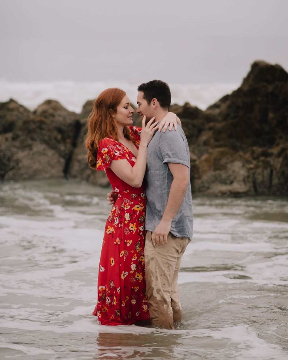 Romantic couple embracing in ocean waves, woman in red floral maxi dress and man in gray shirt and khaki shorts.