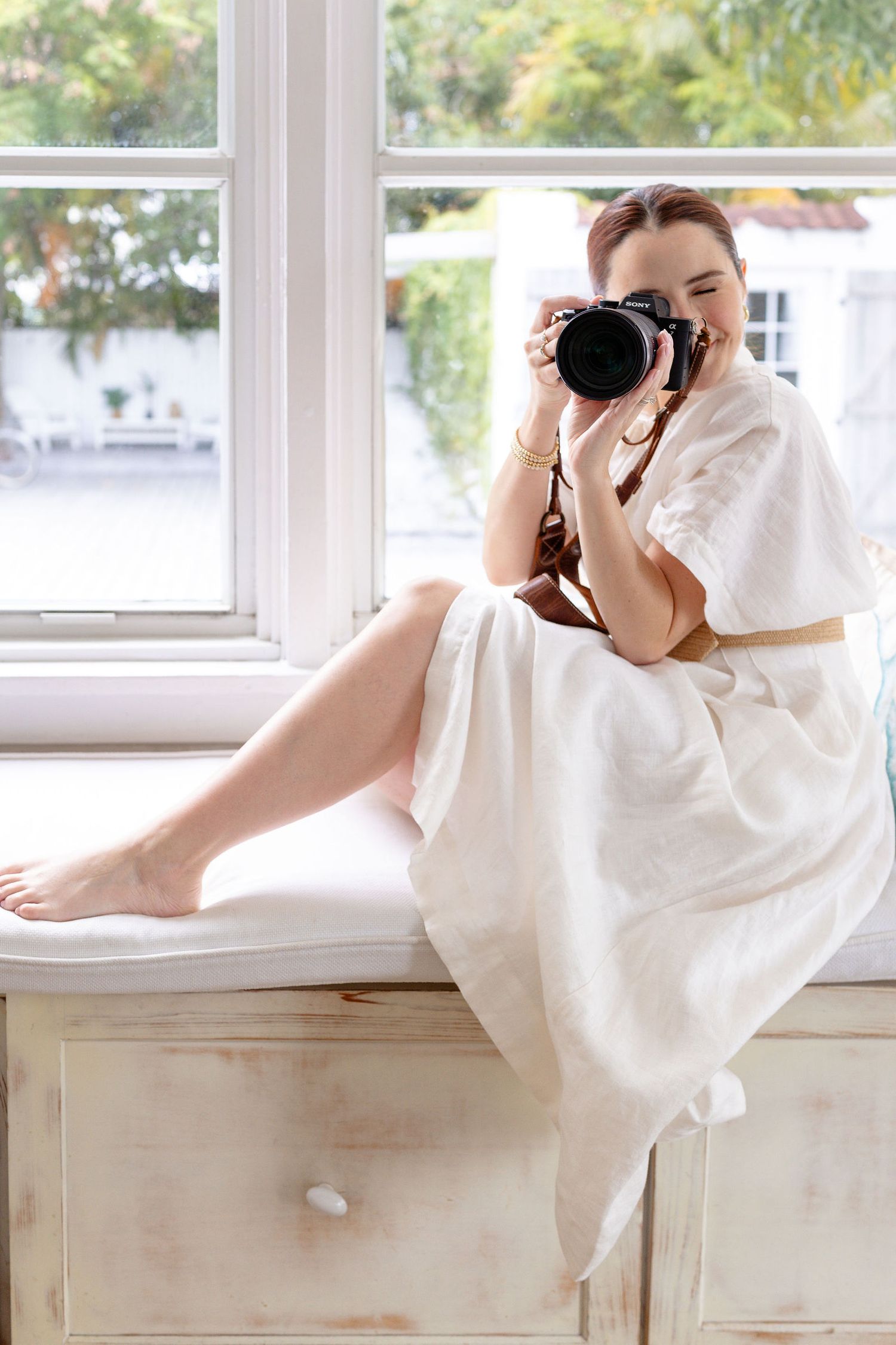 A photographer in a white dress lying on a wooden cabinet while taking photos near a bright window.