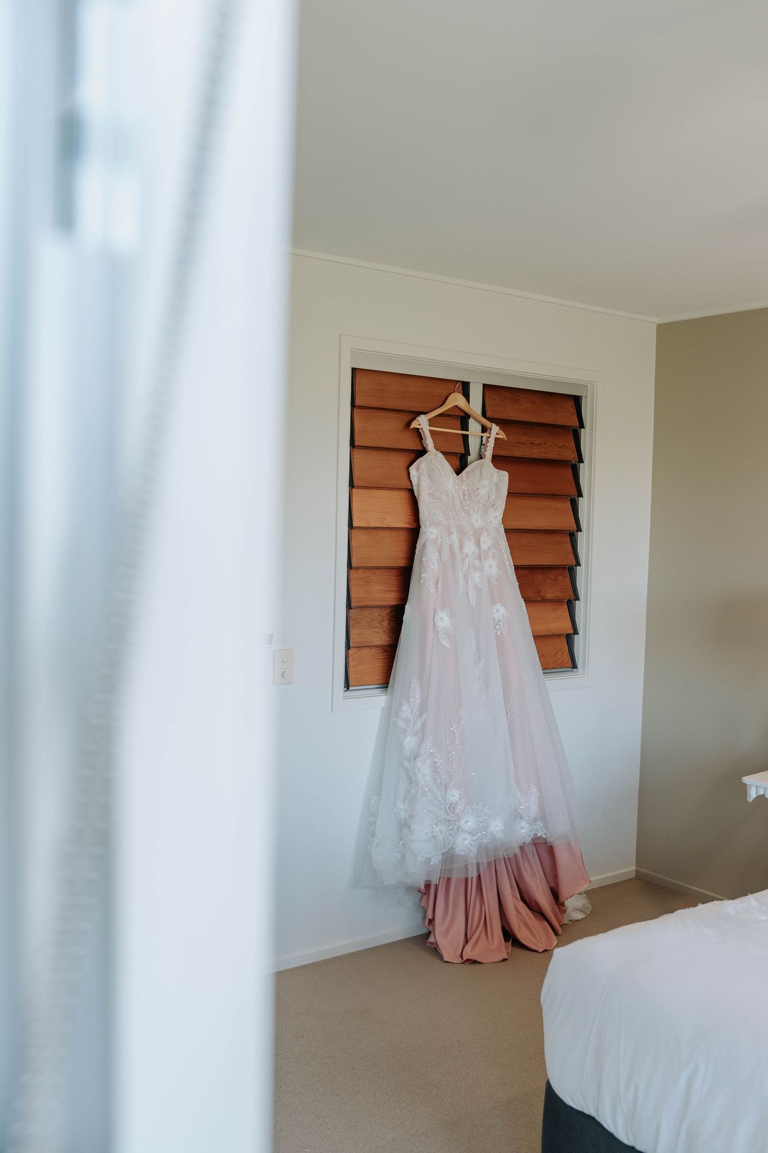 A delicate white lace wedding dress hangs on wooden shutters against a cream wall in a bright bedroom.