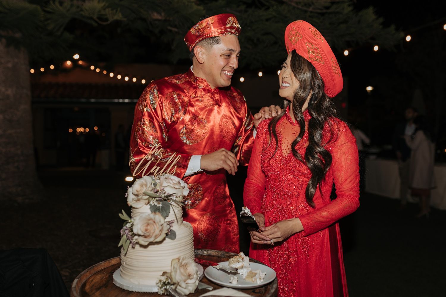 A couple in red Vietnamese attire shares a slice of white wedding cake at an evening reception with string lights in the background.