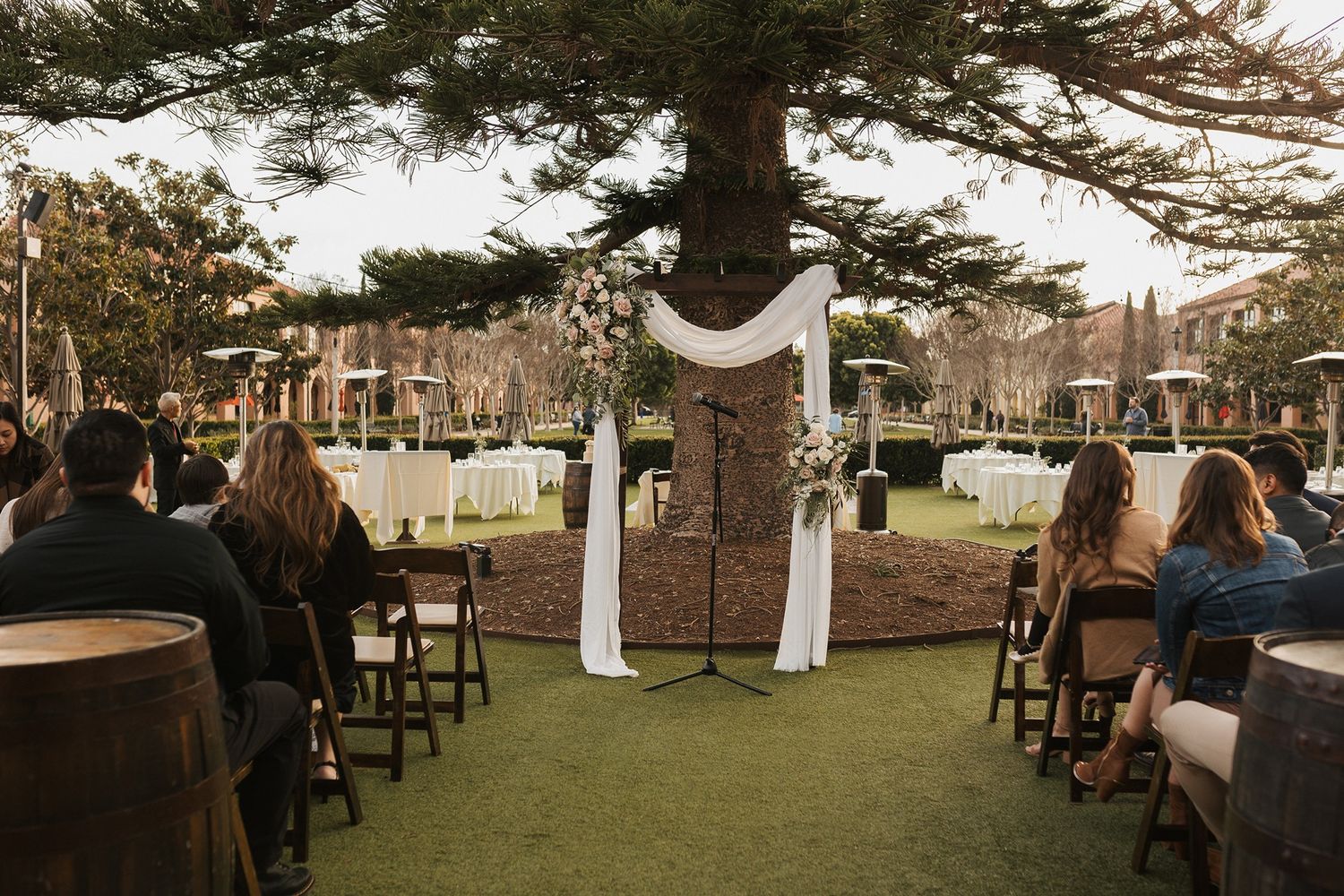 Wedding guests seated in white chairs at an outdoor ceremony venue with tall pine trees in the background at Stone Brewing Liberty Station.
