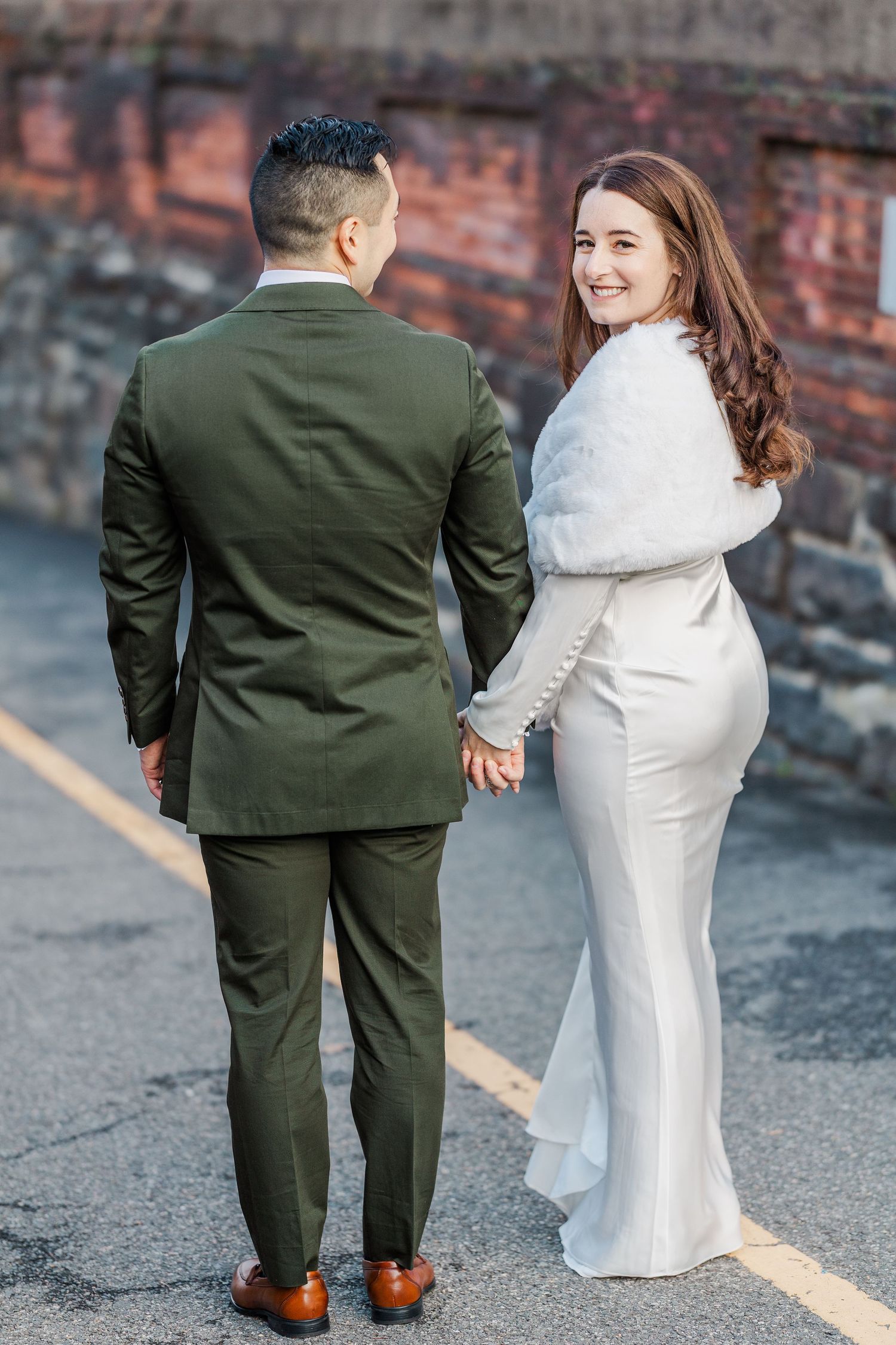 A couple in wedding attire, with the bride in white and groom in dark green suit, photographed from behind on a brick street.