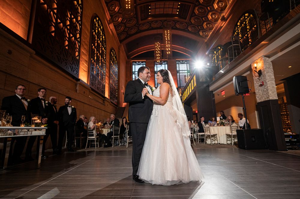 Couple shares their first dance in an elegant ballroom with warm lighting and high ceilings.