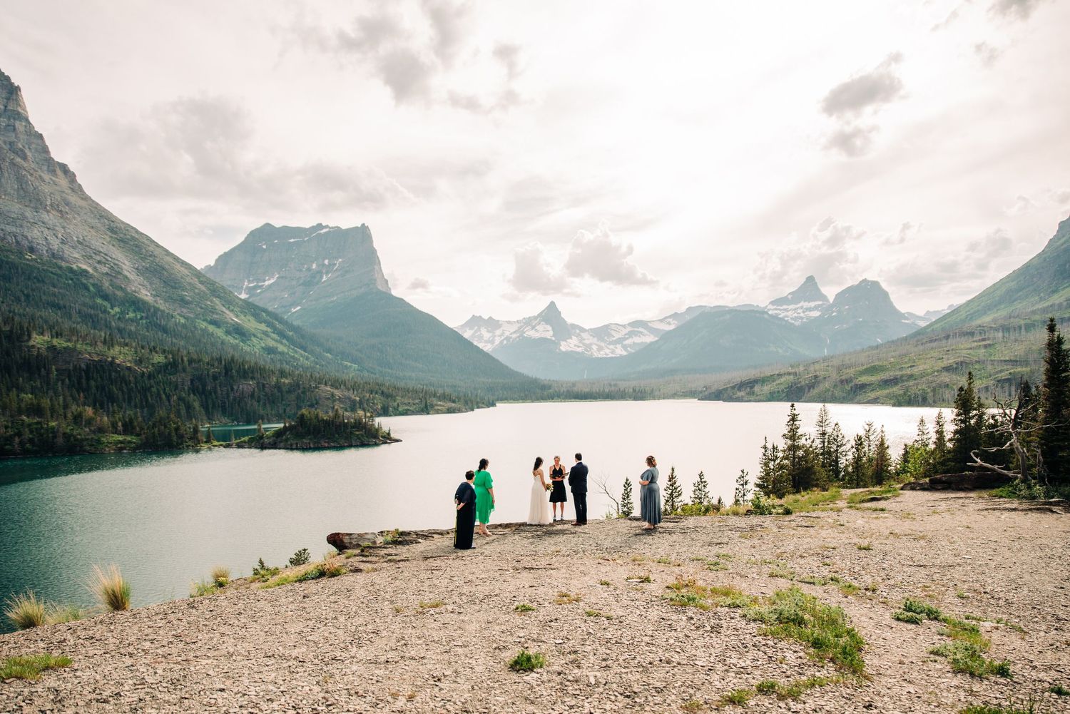 A panoramic view of Glacier National Park shows mountains, lake and shoreline under cloudy skies.