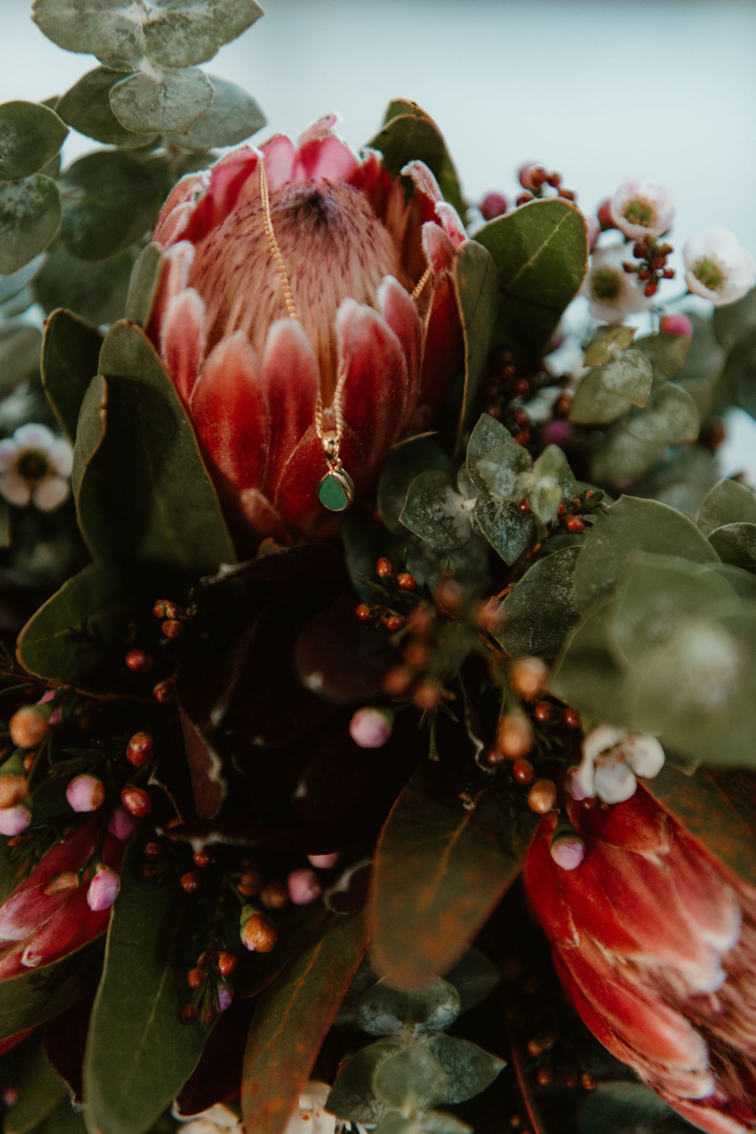 Close-up of a lush bouquet featuring red protea flowers, eucalyptus, berries, and a delicate gold pendant necklace.