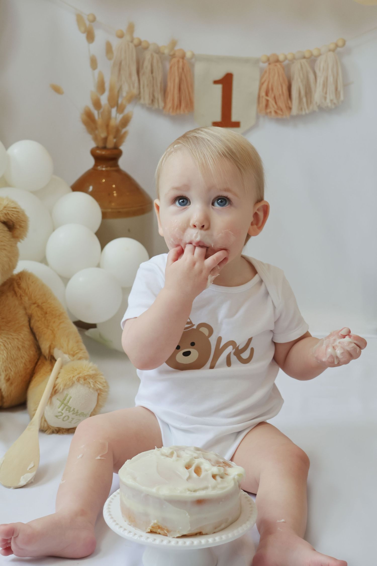 Baby in white onesie sitting on floor with cake and teddy bears during first birthday celebration with decorative banners.