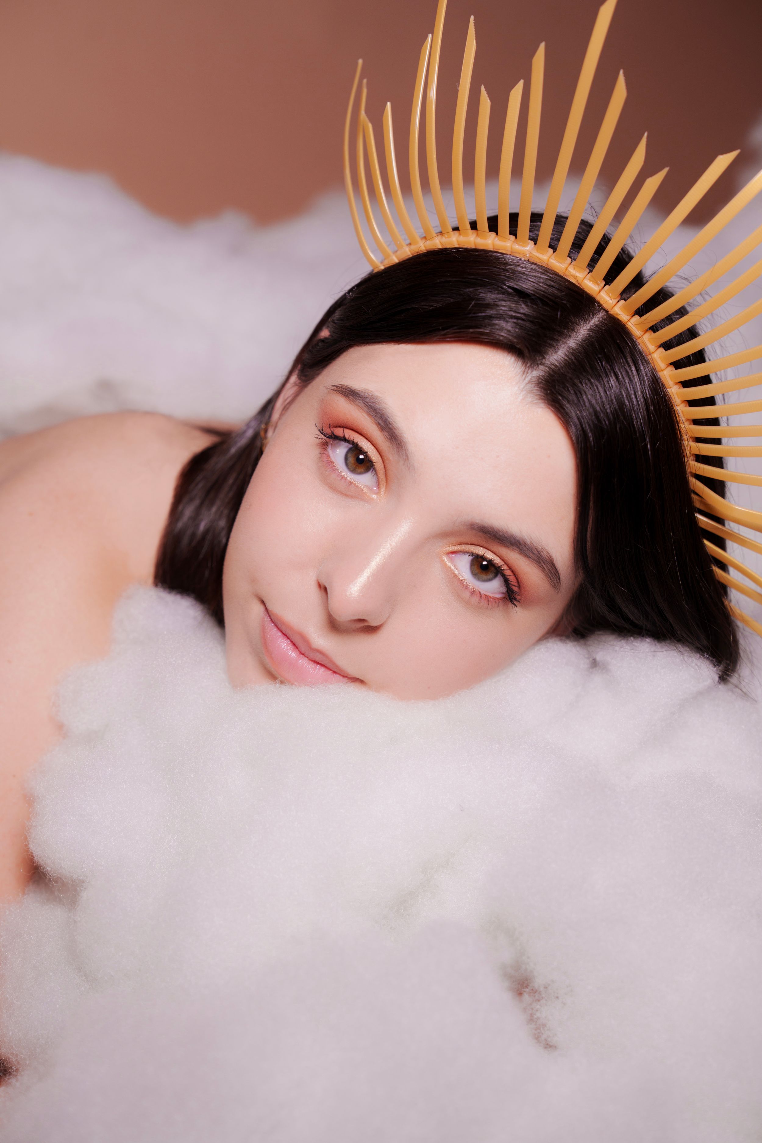 A ethereal fashion portrait with gold spiked crown against soft white clouds and pink backdrop.