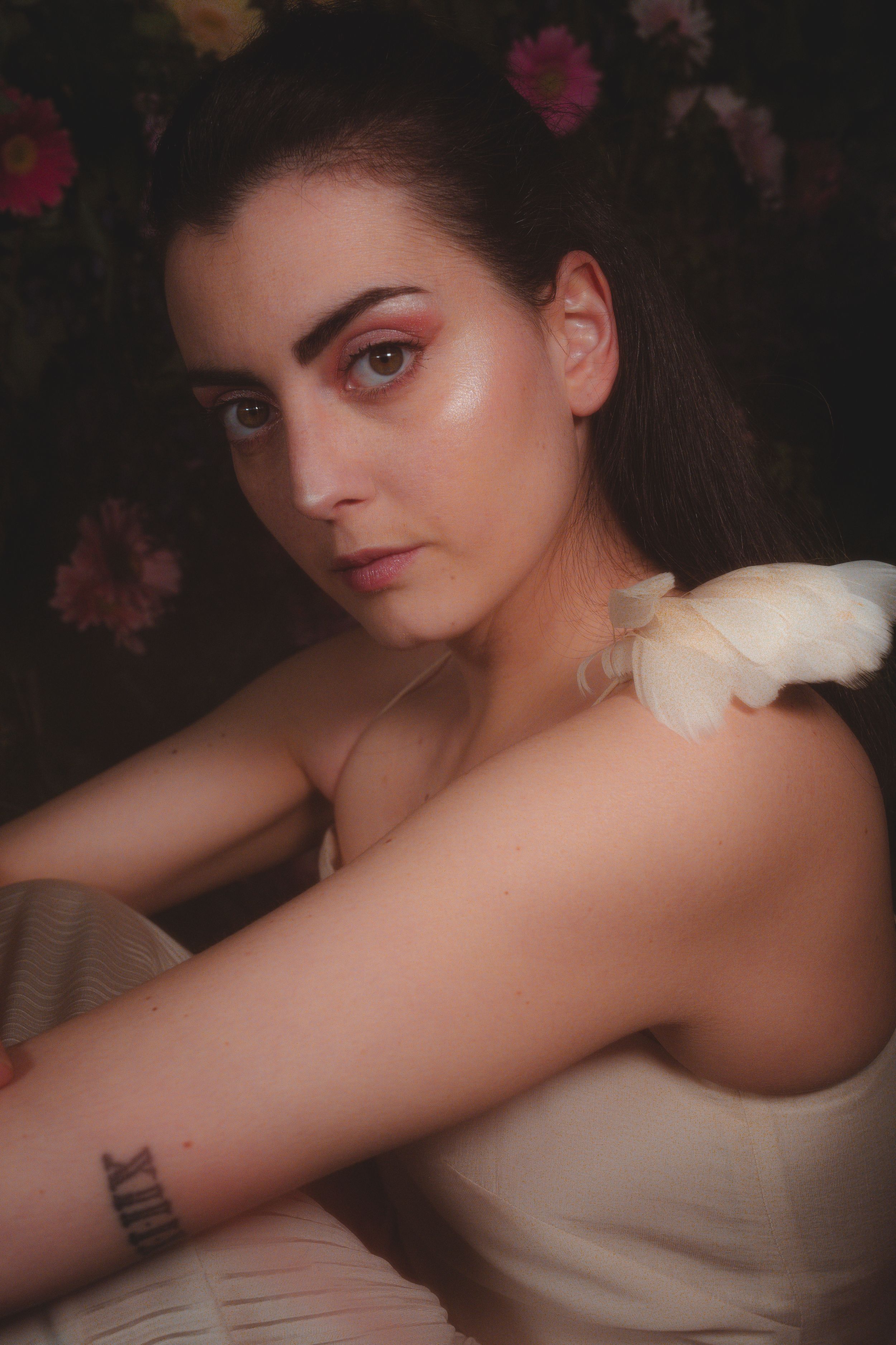 Romantic portrait with dramatic lighting and white feather accent against floral backdrop.