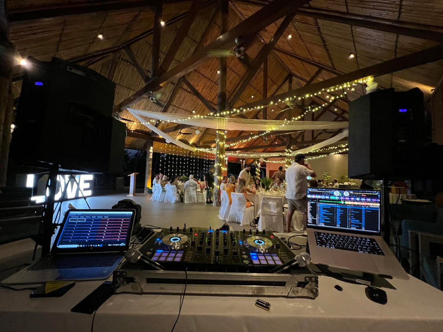 Professional DJ setup with laptops and controllers at a rustic venue decorated with string lights and wooden beams.