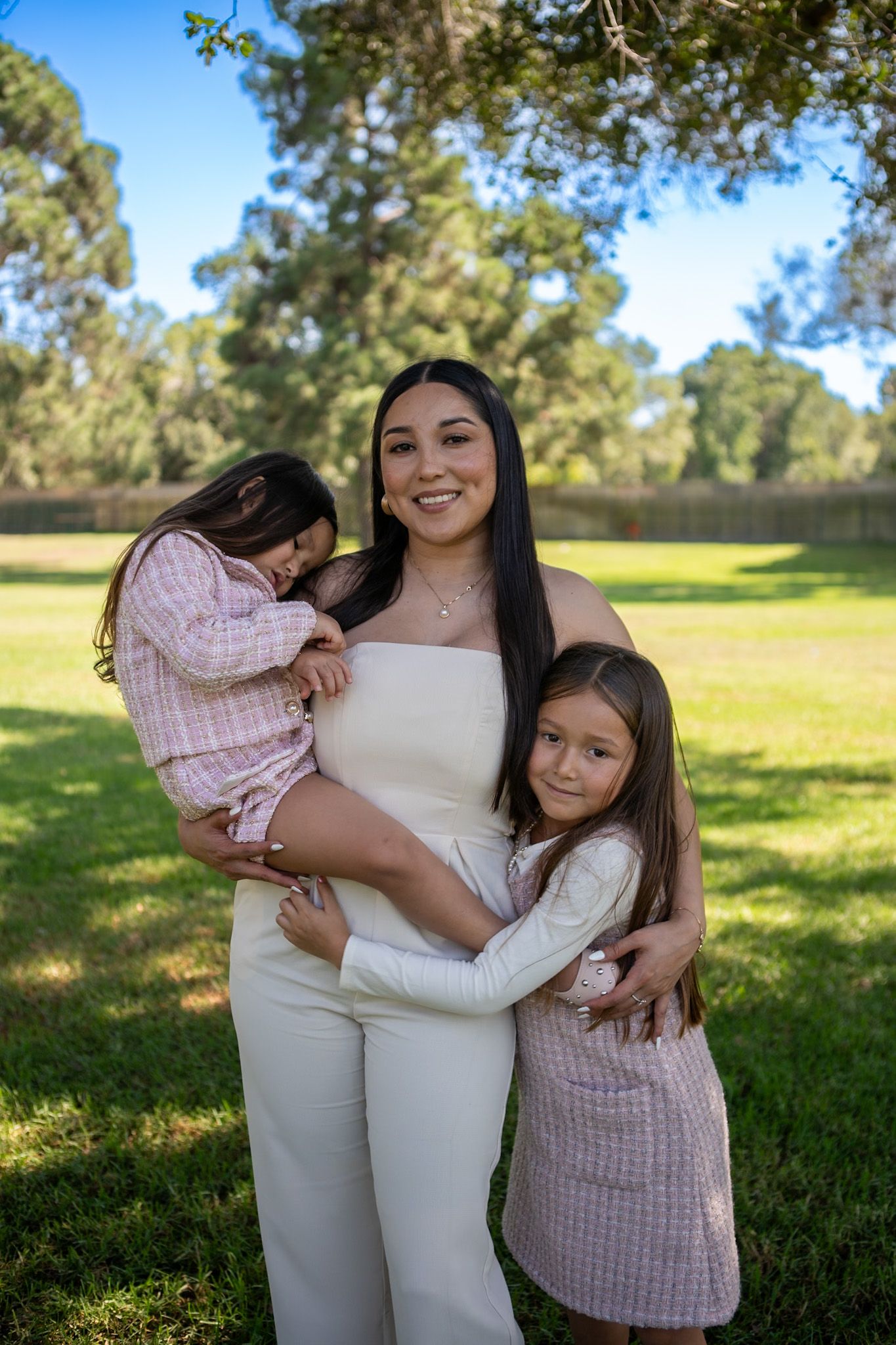 A family embraces while standing in a sunny park with green grass and trees in the background.
