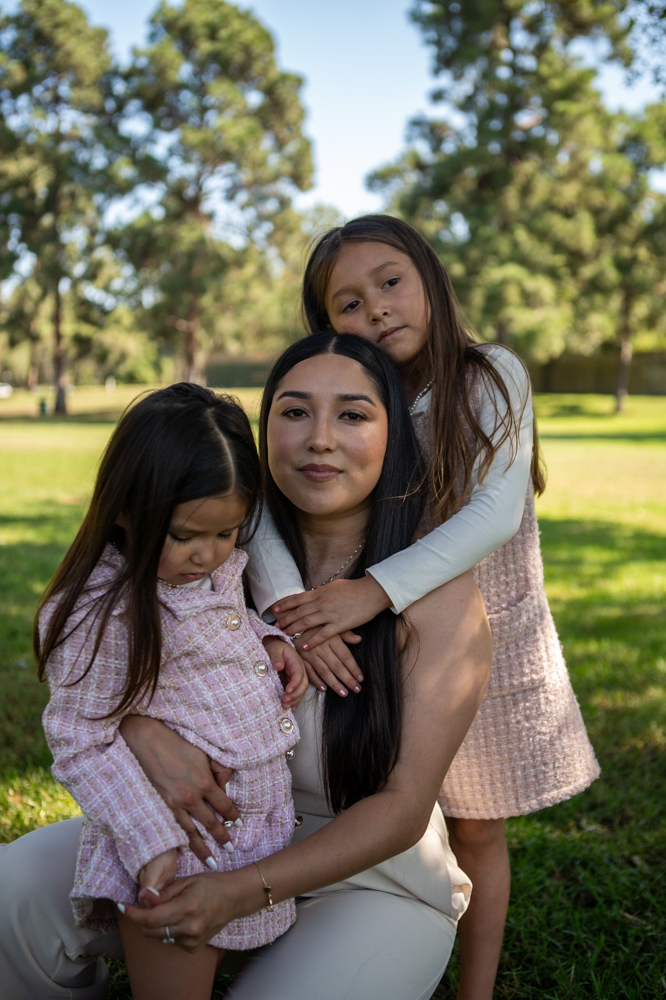 A loving family portrait taken outdoors in a sunny park with green grass and trees in the background.