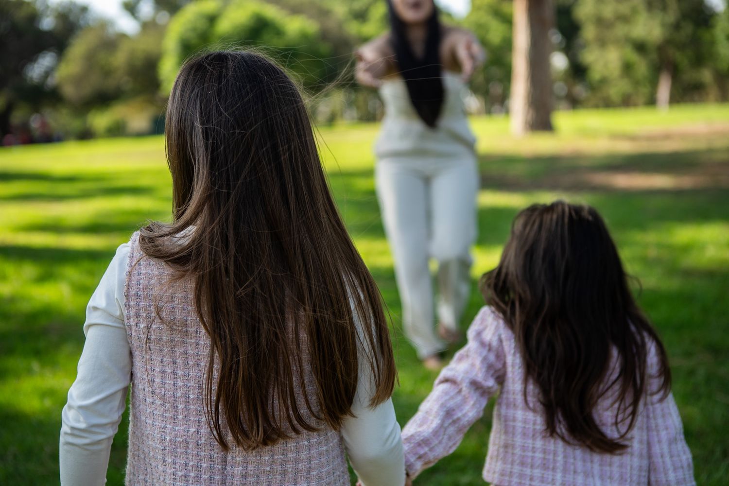 Three people wearing white and pink sweaters walk together in a sunny park setting.