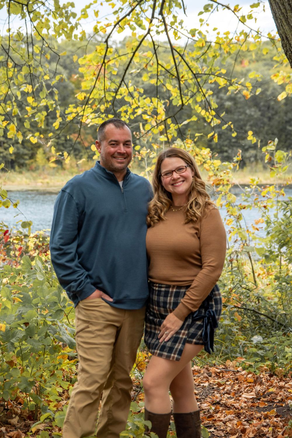A couple poses together among fall foliage near a lake during autumn.
