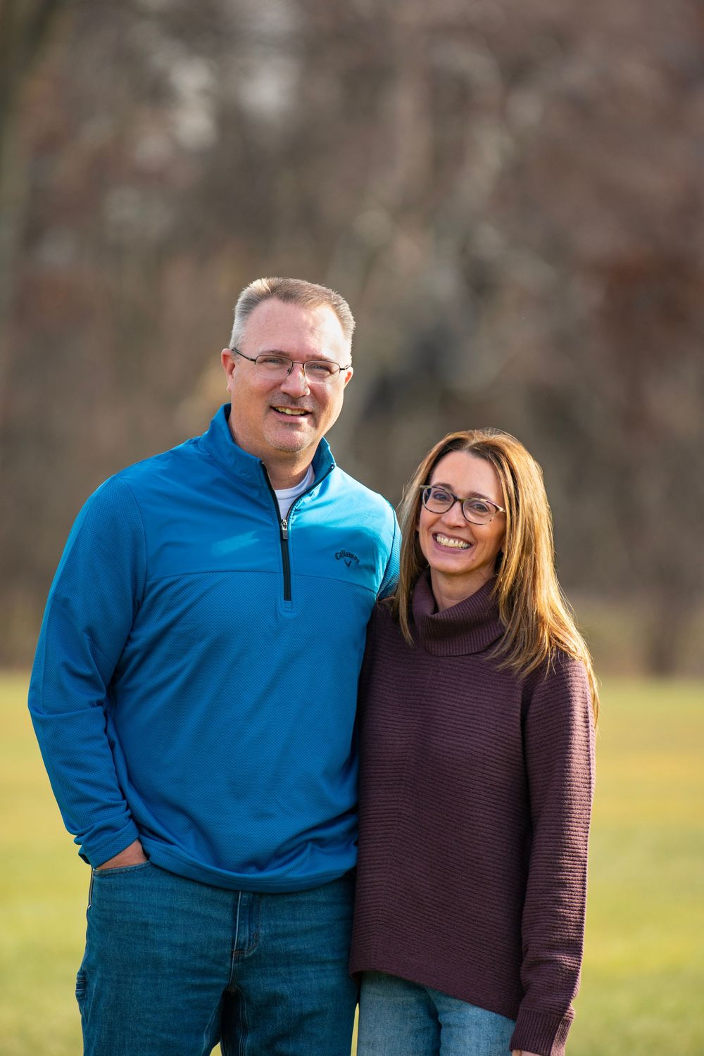 A couple smiles warmly while posing together outdoors in casual sweaters.