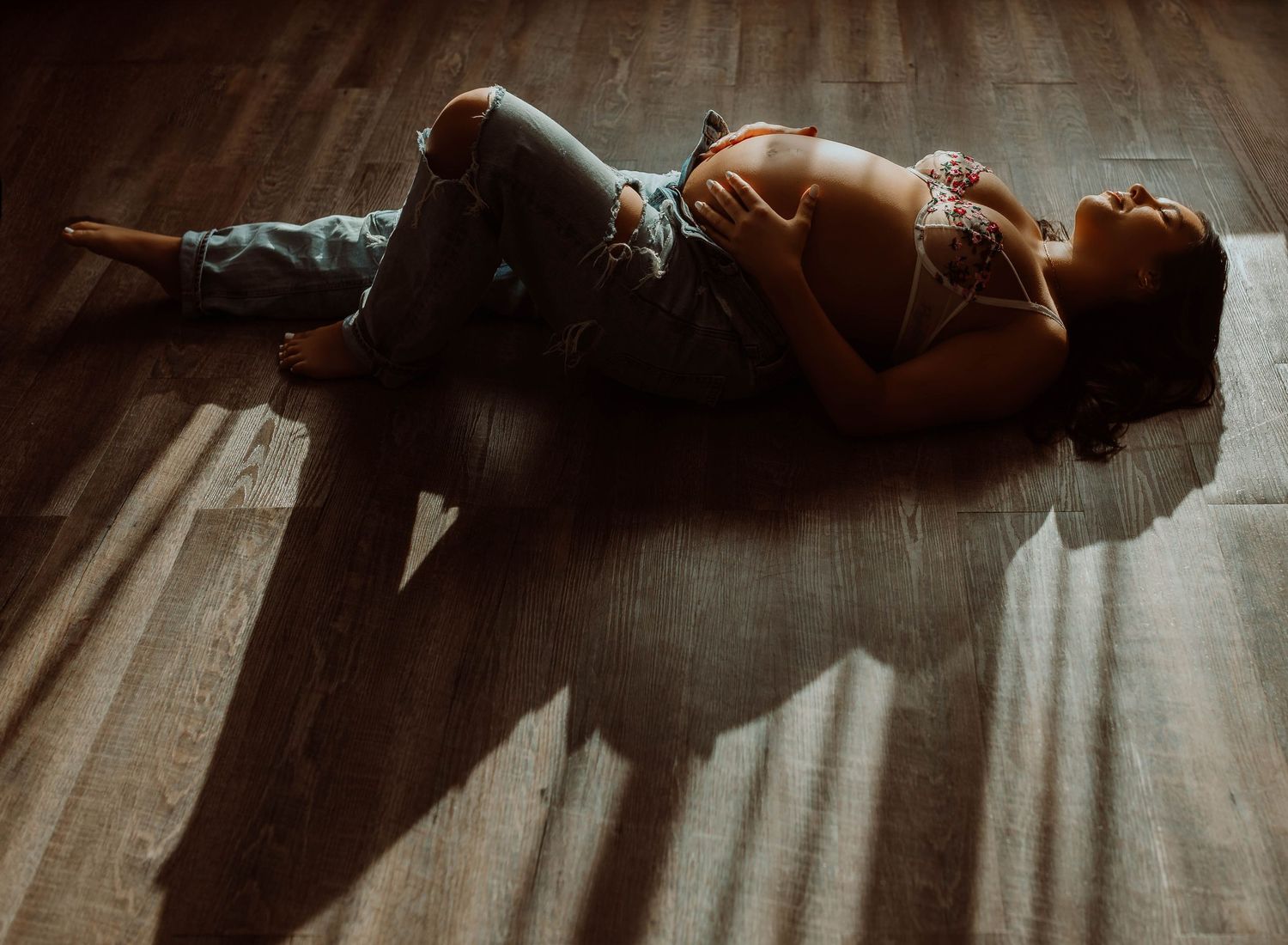 Stillwater Minnesota boudoir, pregnant woman lying on hardwood floor in natural light, wearing jeans and a crop top, holding her baby bump.