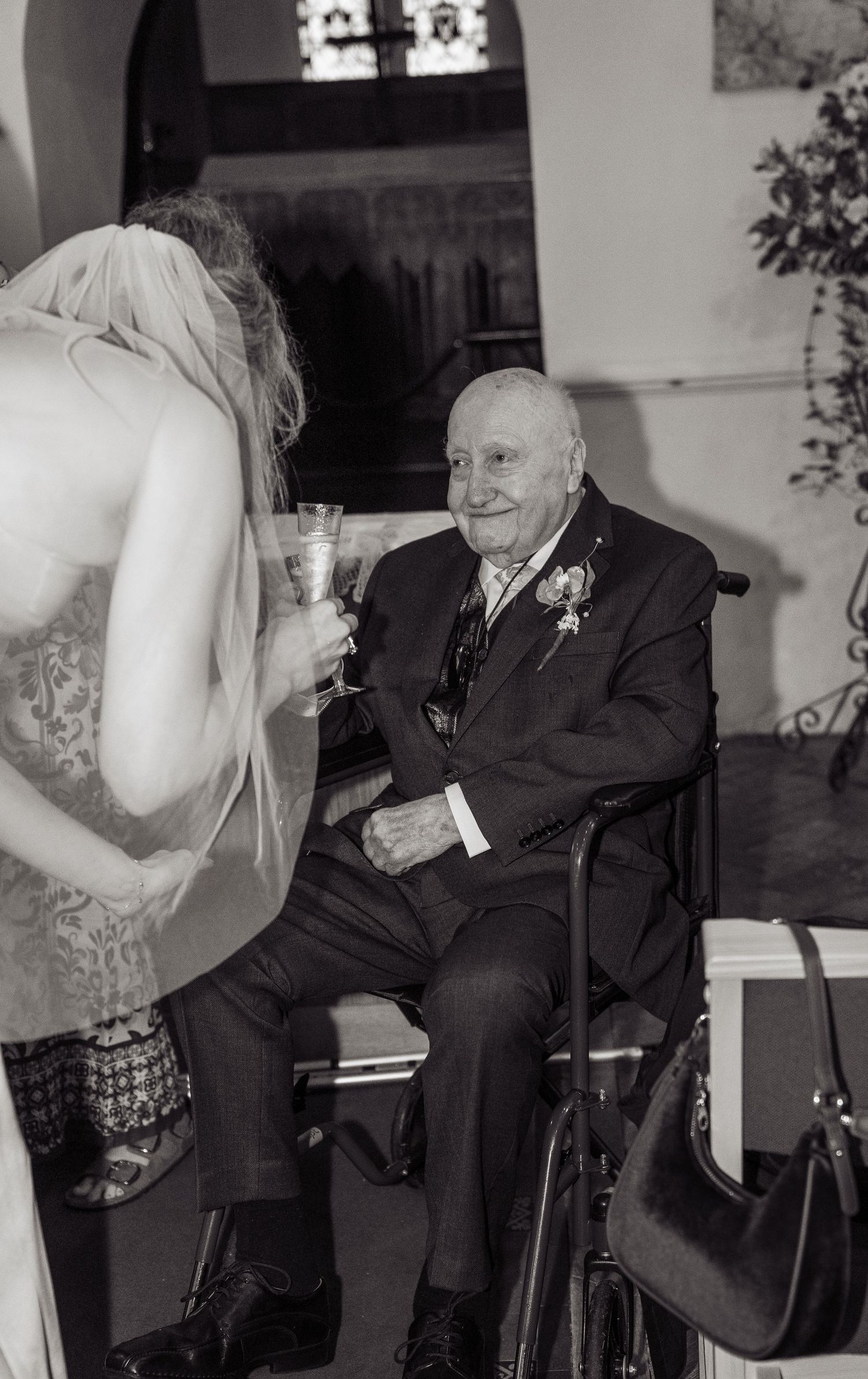 Elderly man in wheelchair smiling at bride holding champagne glass during wedding reception, black and white photo.