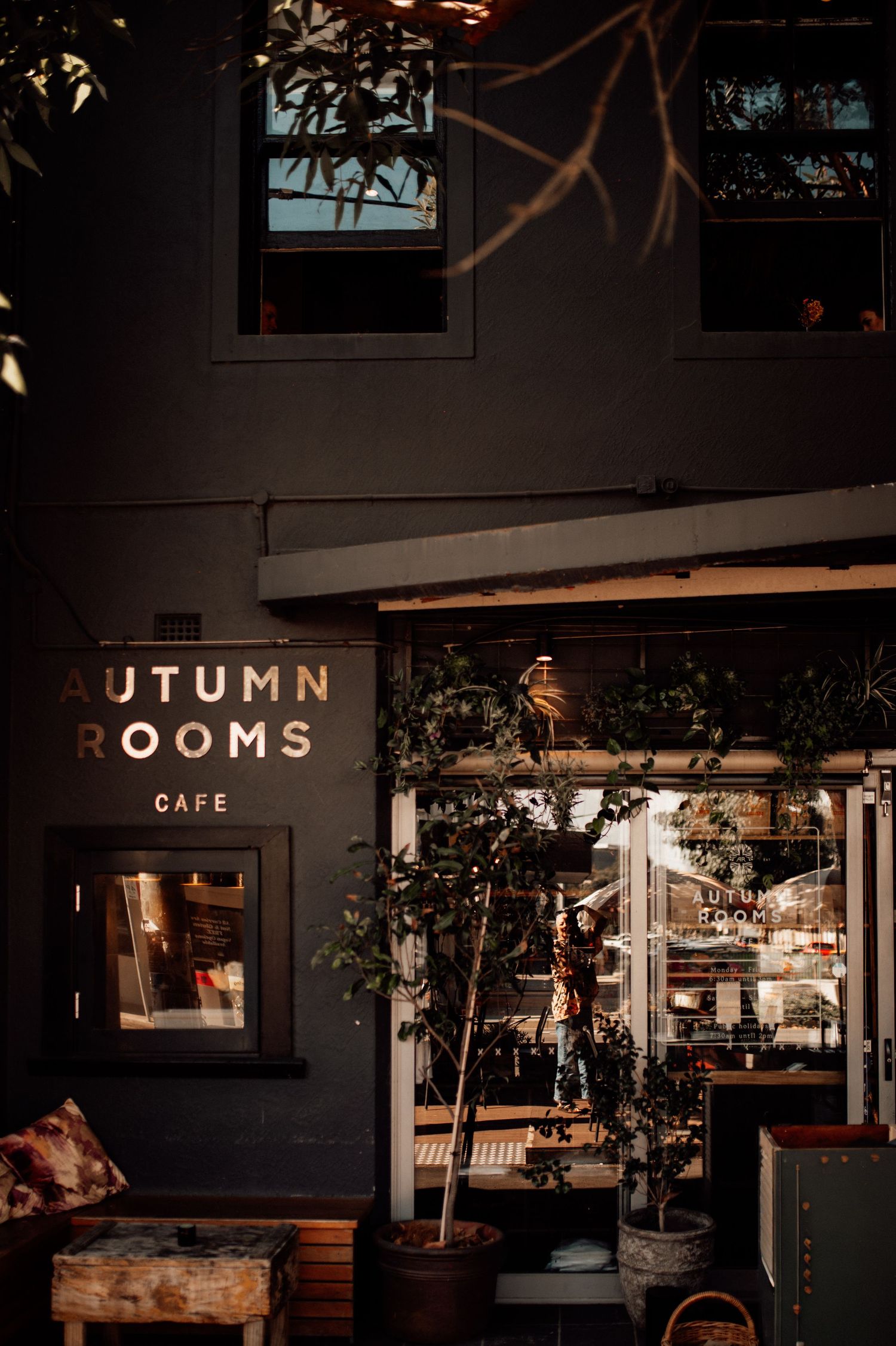 Autumn Rooms Cafe exterior at dusk with warm glowing signage, potted plants, and rustic wooden furniture on a dark facade.