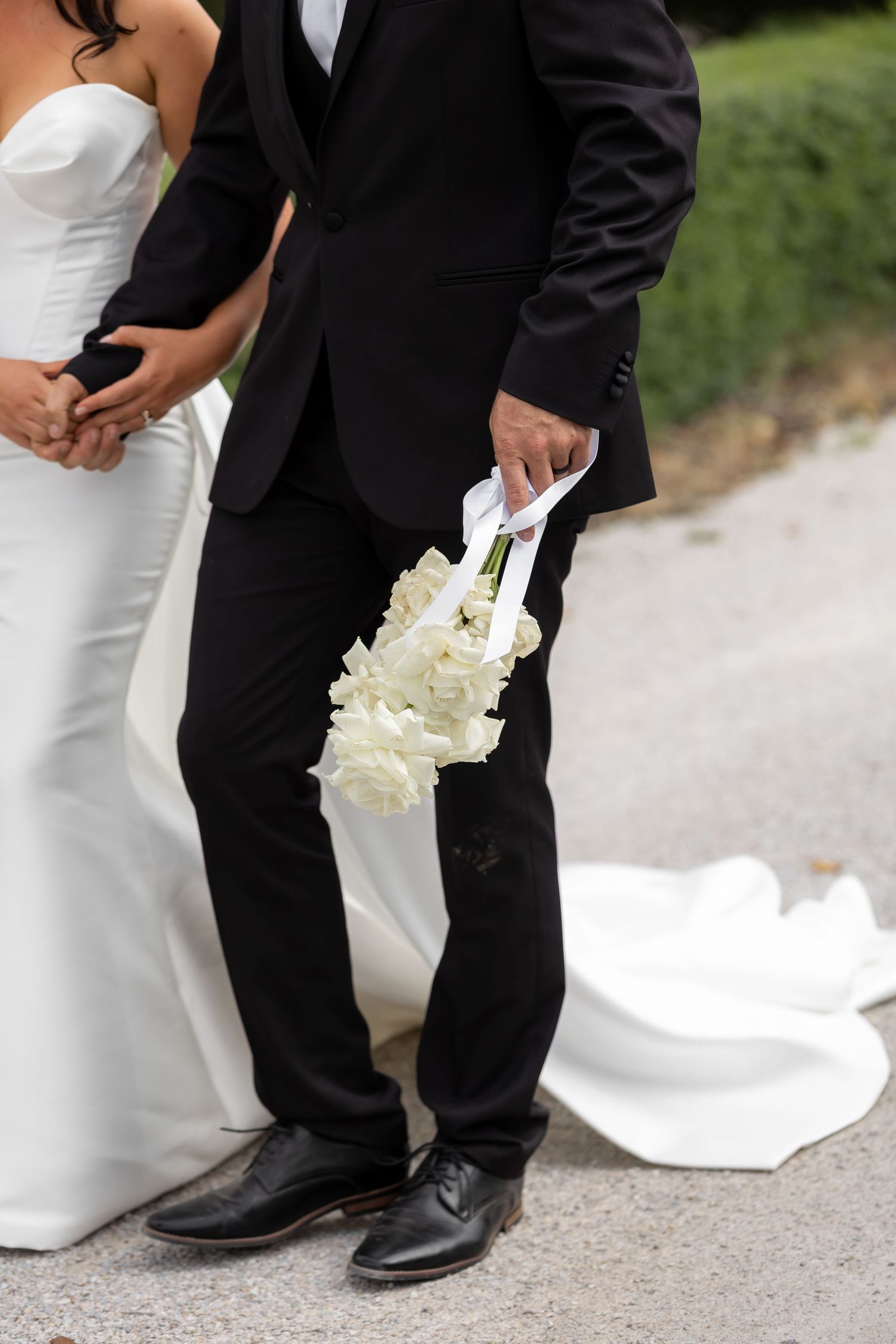 Groom in black suit holding white rose bouquet with ribbon beside bride in white wedding gown outdoors.