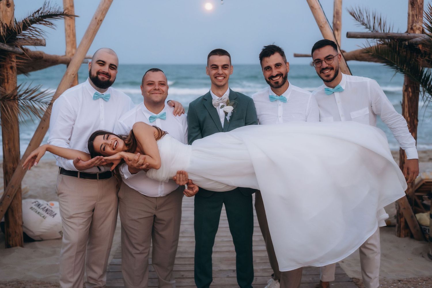 Groom holds bride while groomsmen pose together on a beach wedding celebration at sunset near the ocean.