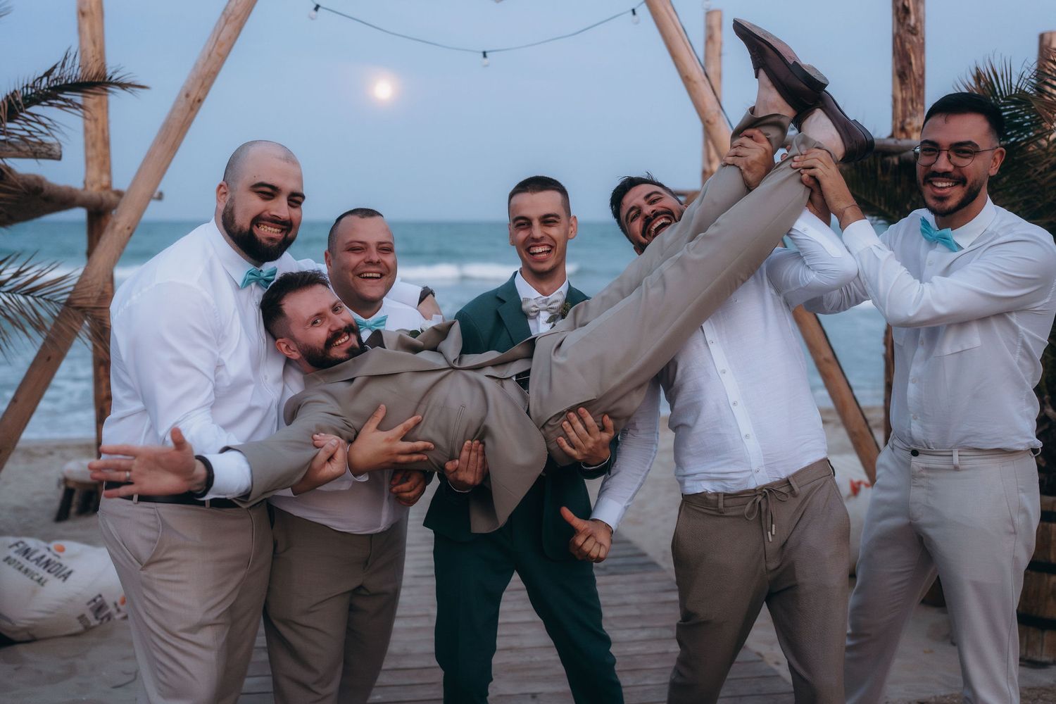 Groomsmen lifting groom on beach wedding celebration at dusk with wooden arch and ocean backdrop.