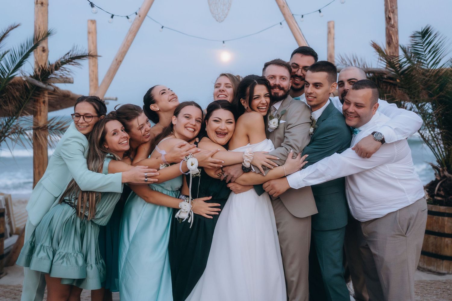 Wedding party group hug on beach at dusk, bride in white gown surrounded by bridesmaids in teal dresses and groomsmen.