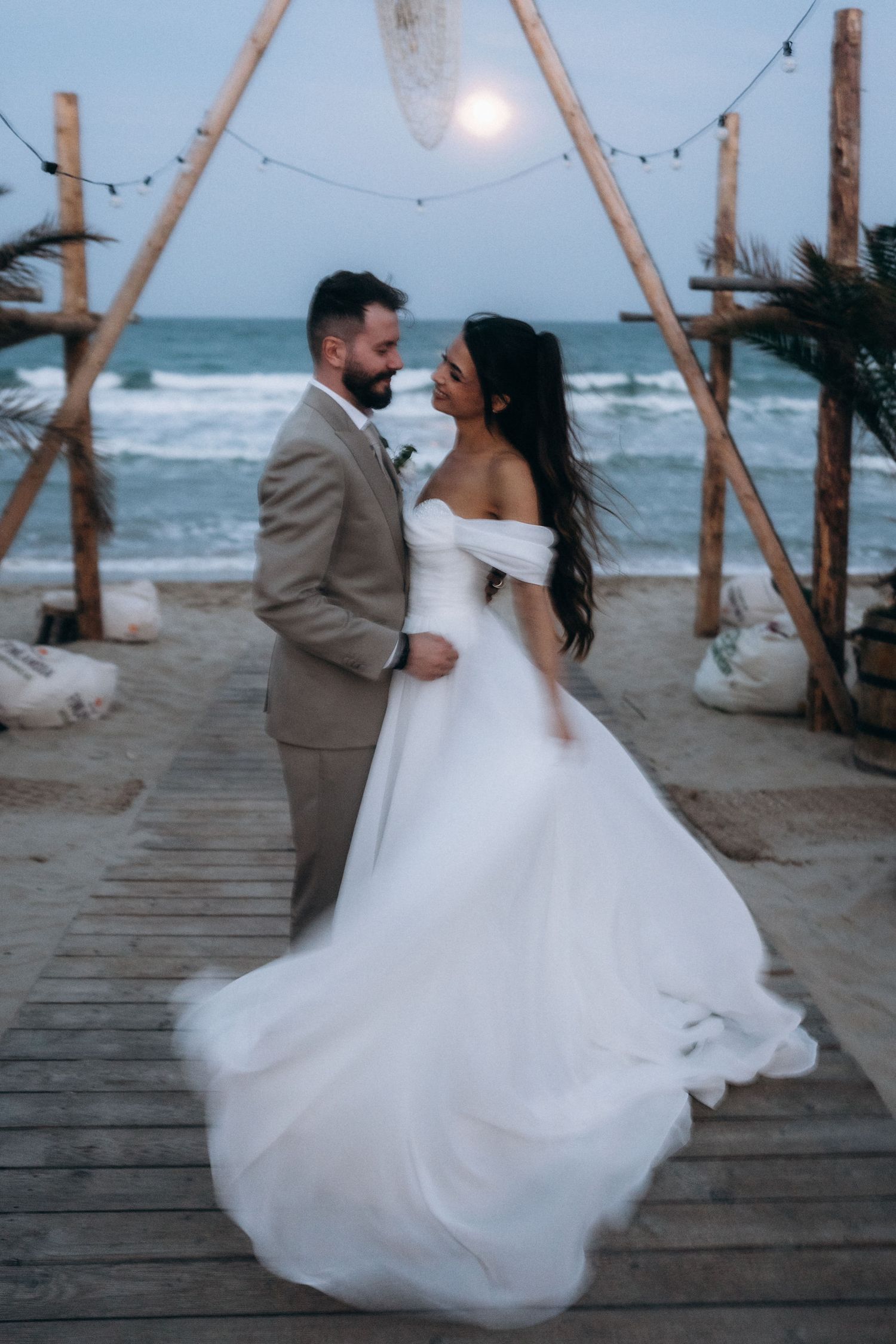 Bride and groom share a romantic moment on a beach boardwalk at dusk, with ocean waves and string lights behind them.