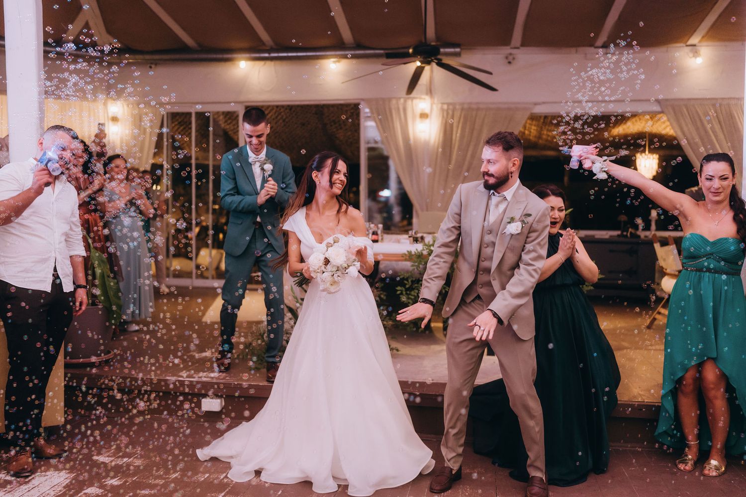 Bride and groom walk through confetti shower surrounded by smiling wedding guests during outdoor reception celebration.