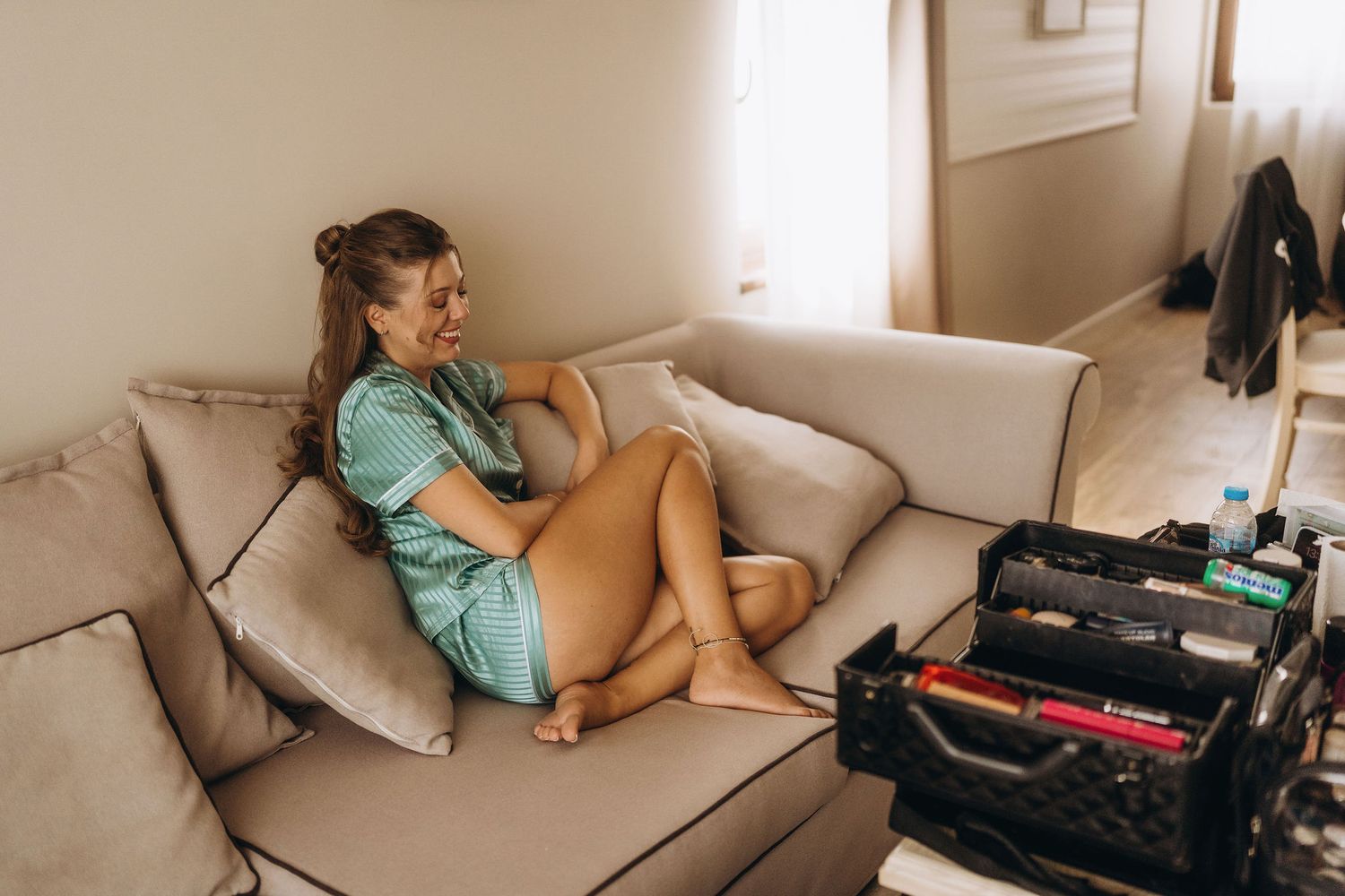 Young woman in teal dress relaxing on beige sofa, smiling while sitting cross-legged near a storage crate indoors.