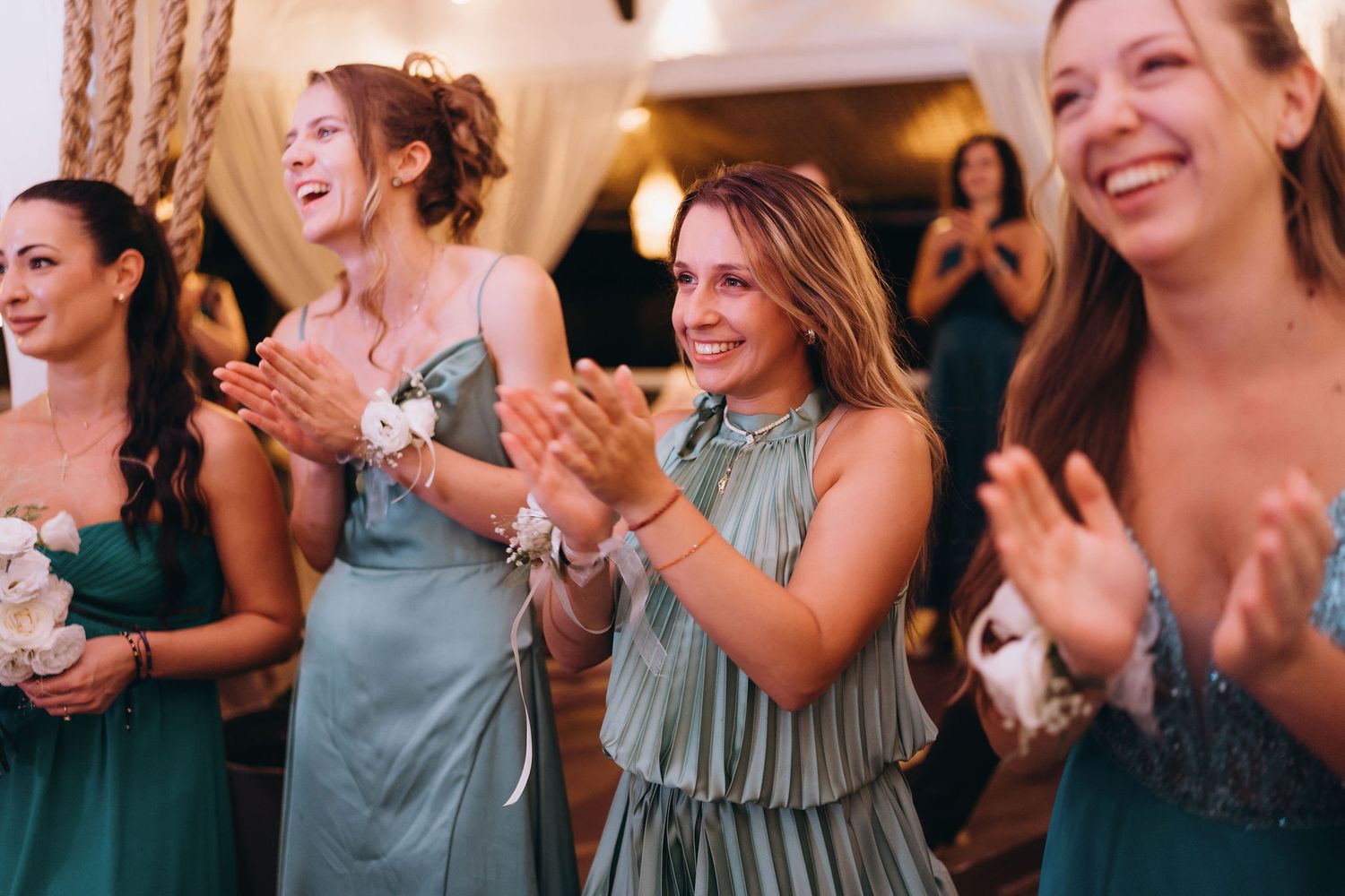 Bridesmaids in teal dresses clapping and smiling joyfully during wedding reception ceremony indoors.