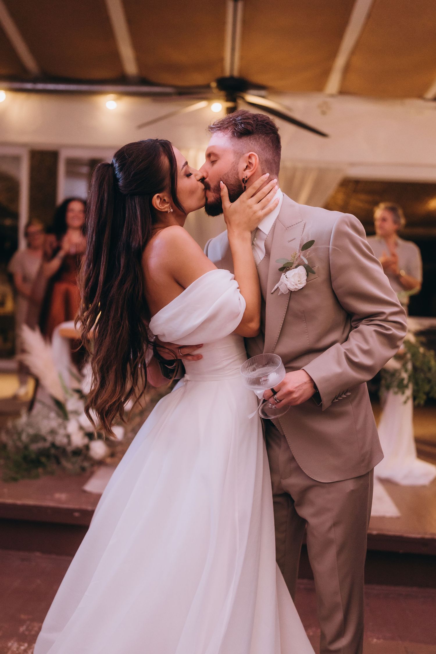 Bride and groom share a romantic first kiss at their wedding reception, with guests celebrating in the background.