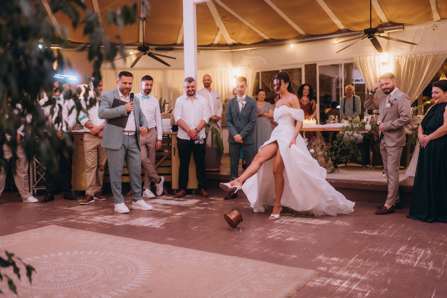 Bride in white wedding dress kicking a football on the dance floor surrounded by guests at an indoor wedding reception.