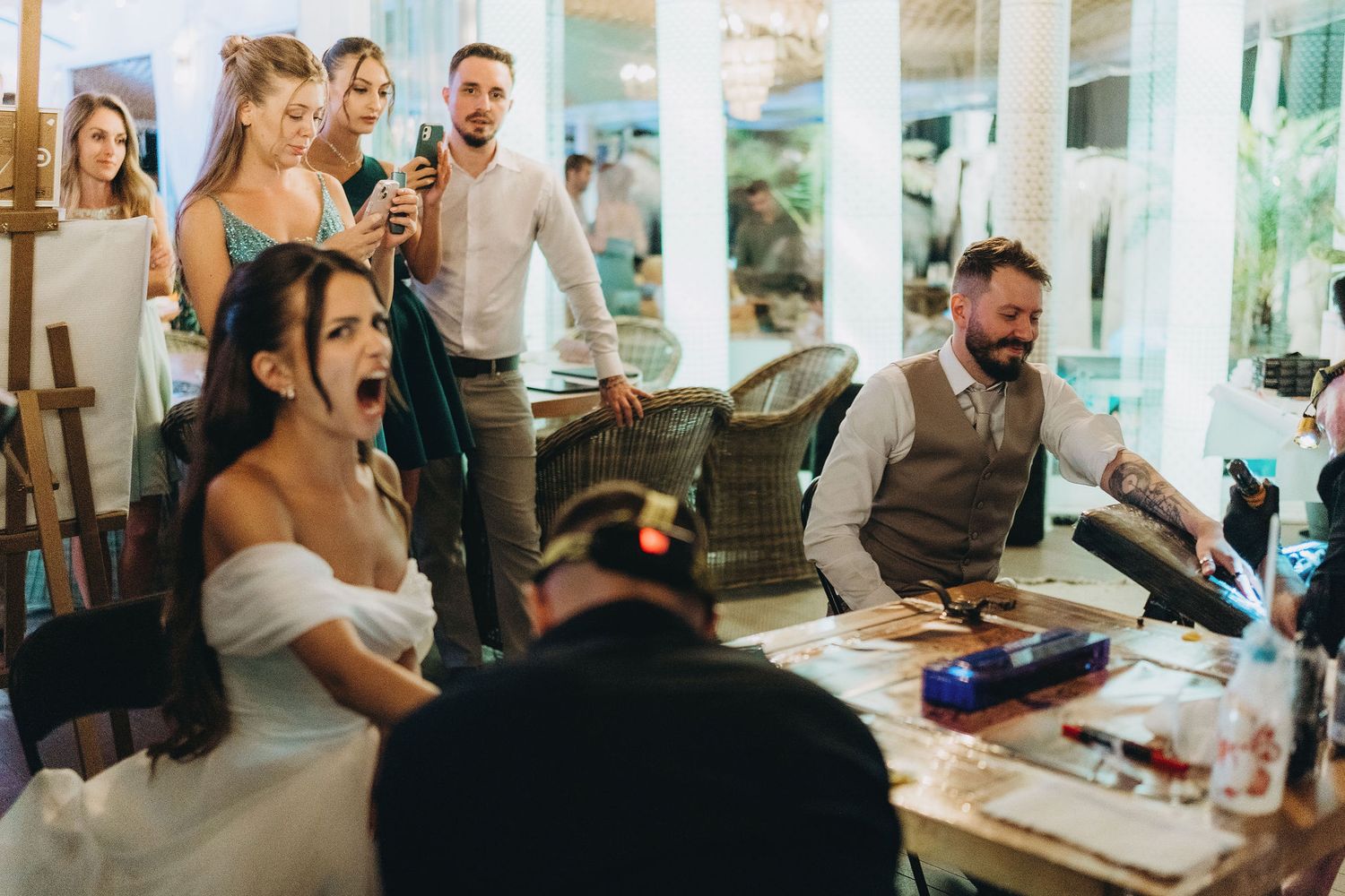 Bride reacts with shock at wedding reception while guests watch and a man sits at a DJ table.
