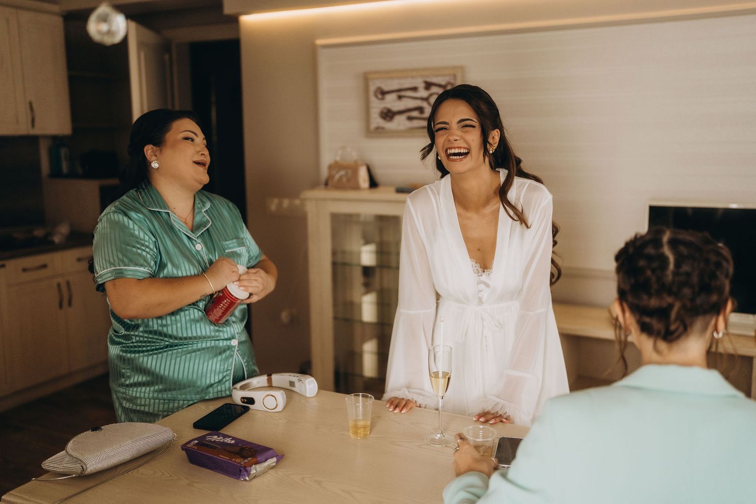 Three women laughing and celebrating together indoors, with champagne glasses, wearing robes in a bright, cozy setting.