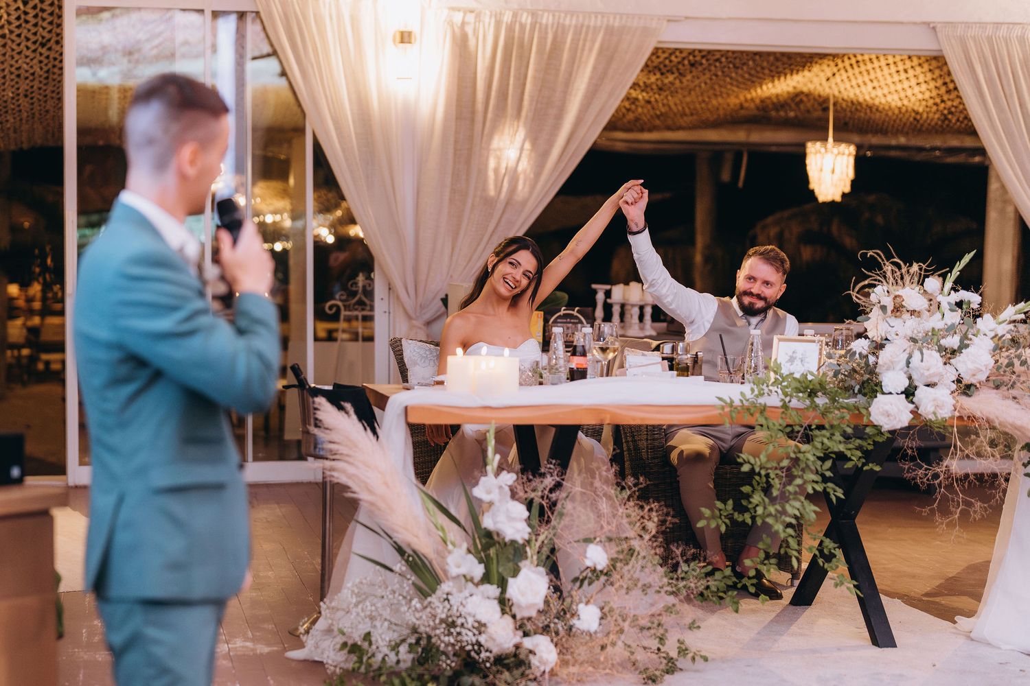 Bride and groom raising hands joyfully at wedding reception table while guest gives a toast.