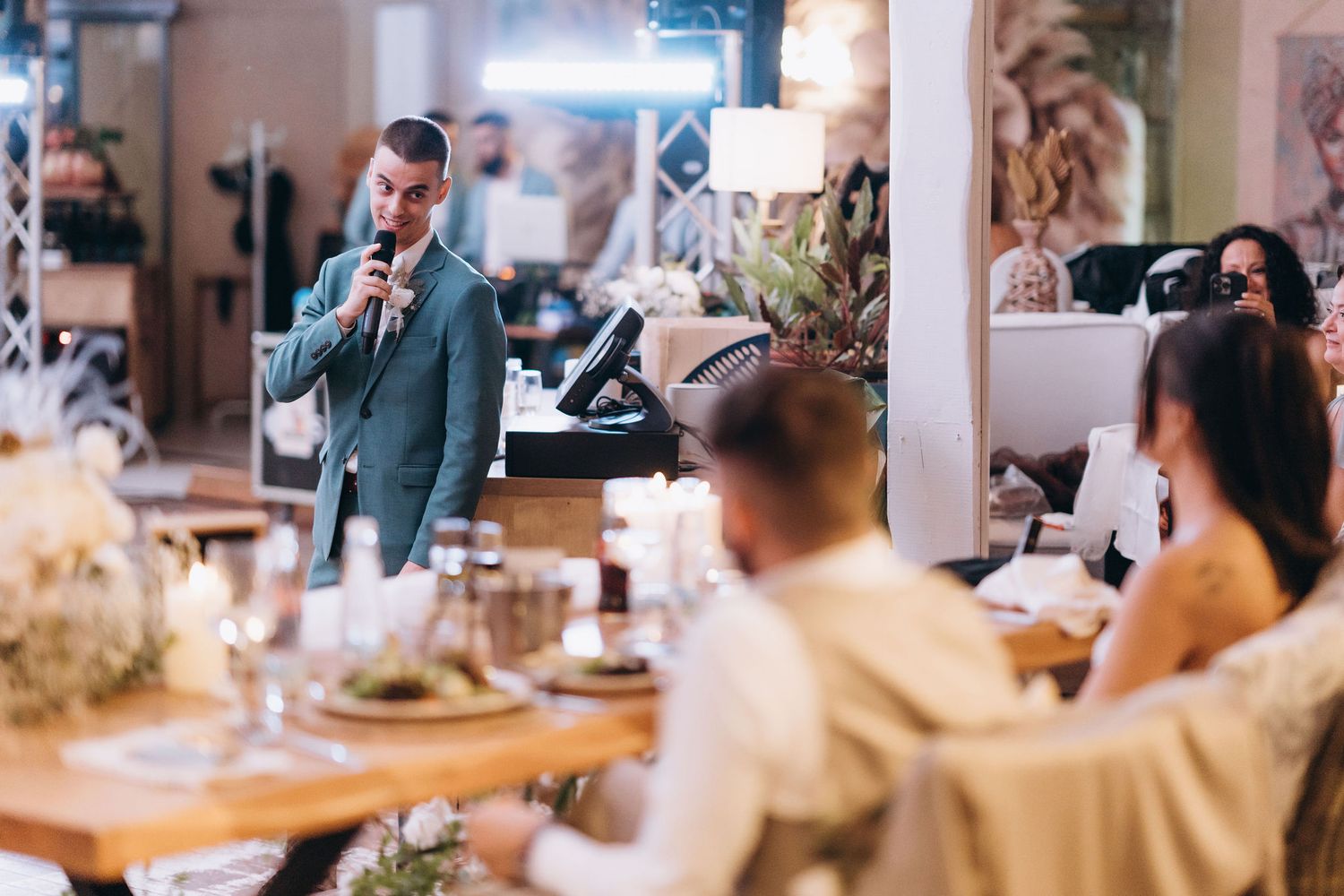 Man in blue suit giving a speech at an elegant wedding reception dinner with candles and floral centerpieces.