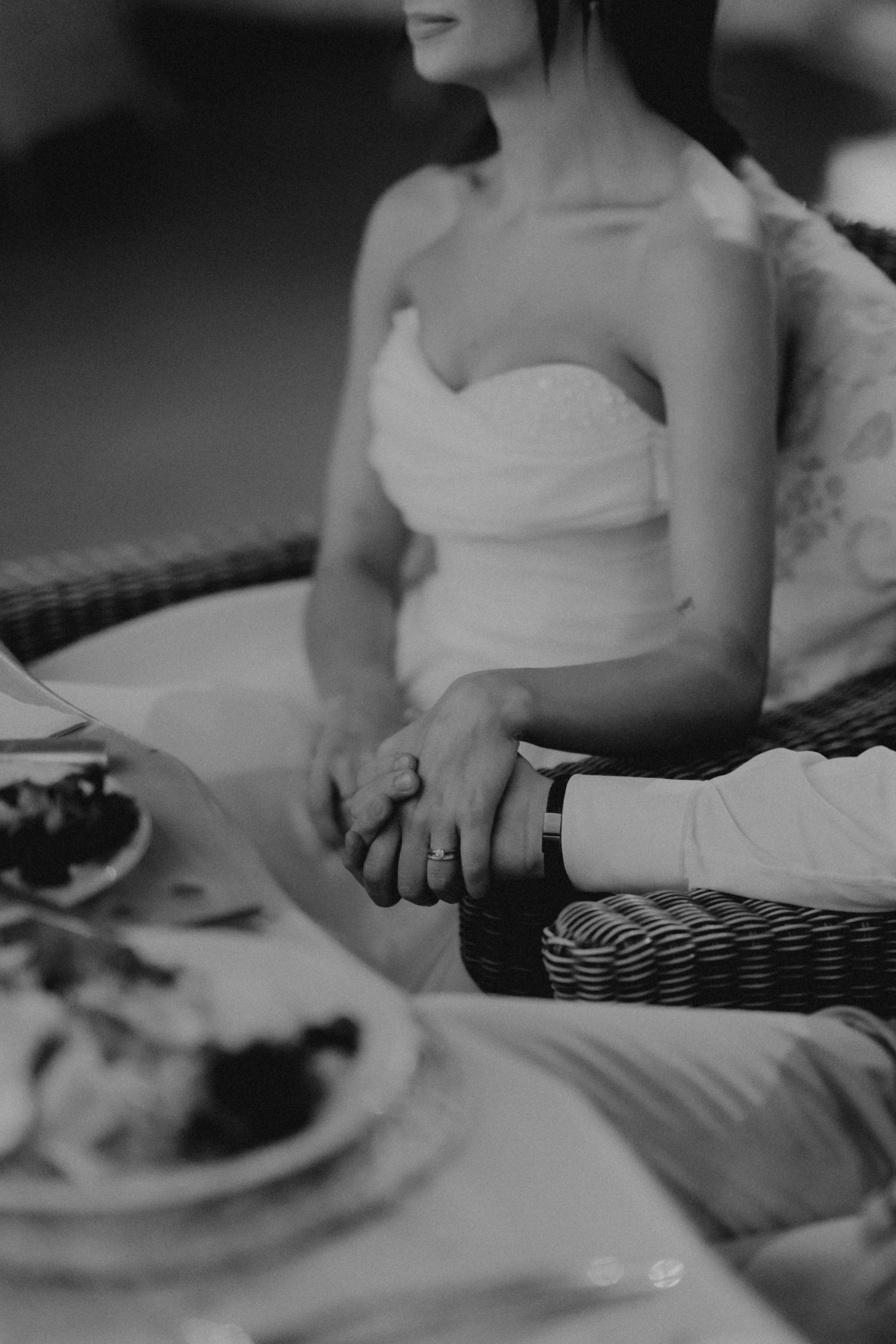 Black and white photo of a bride and groom holding hands at a wedding reception table with food.
