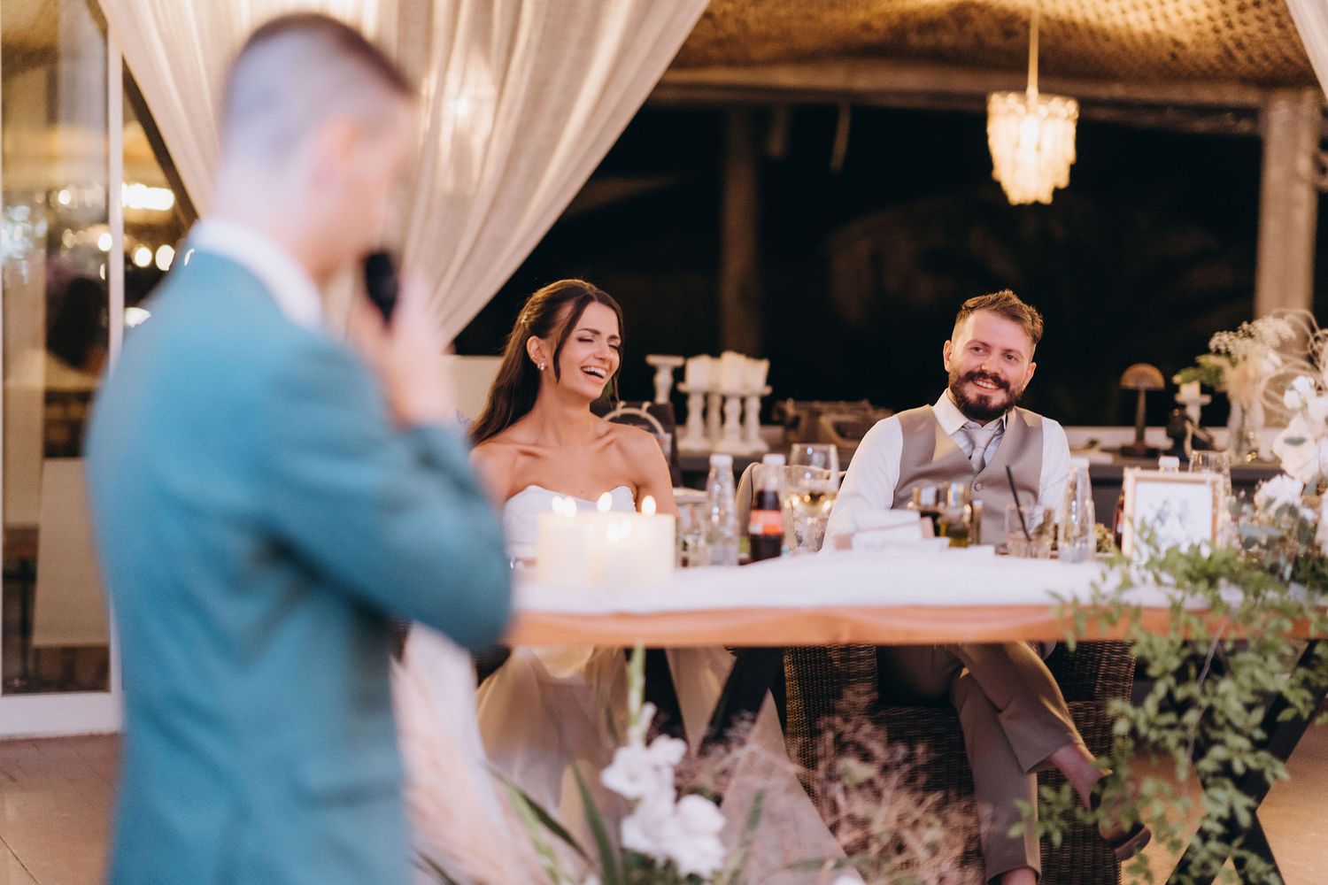 Bride and groom smiling at sweetheart table during elegant indoor wedding reception with candles.