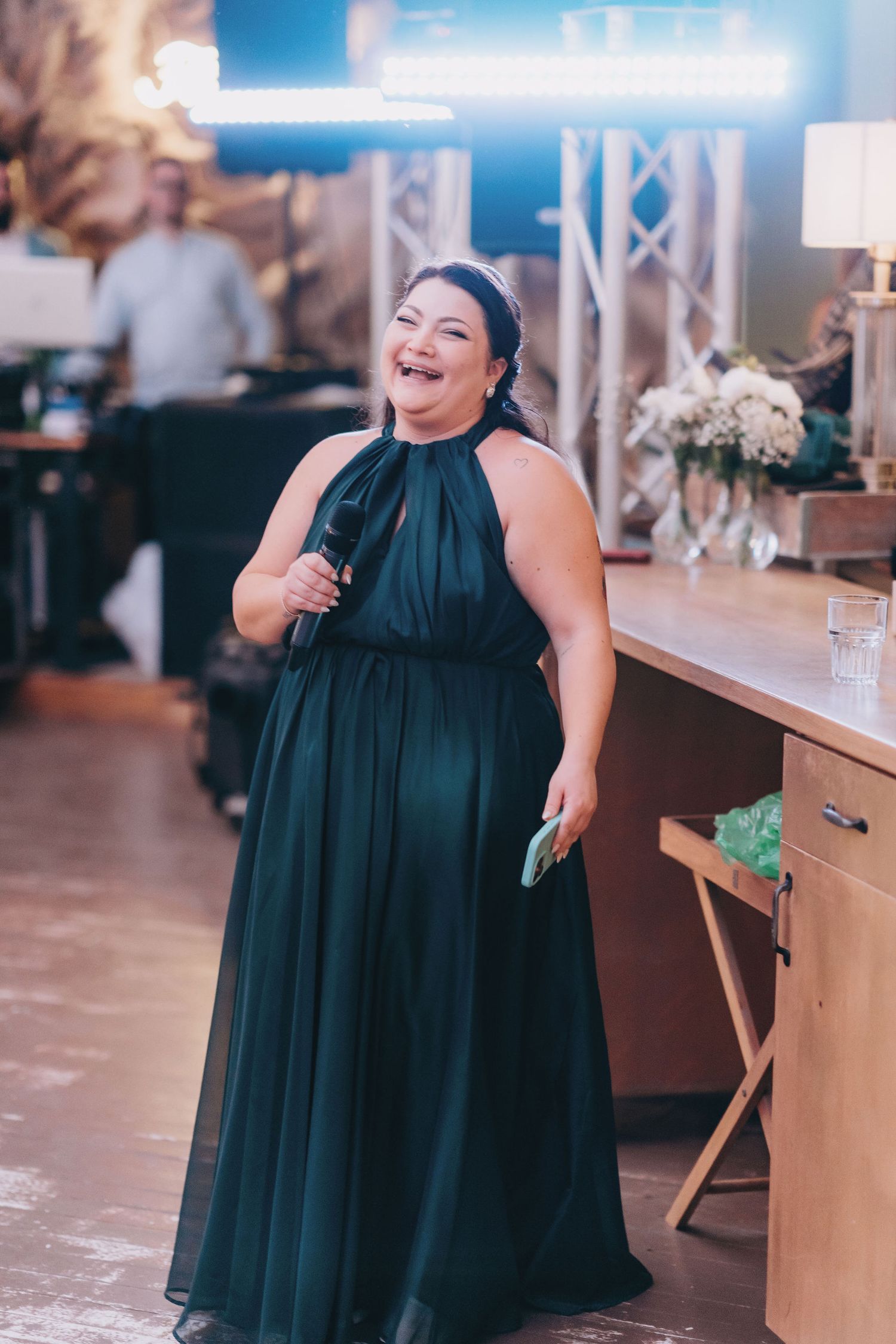 Woman in elegant dark green halter gown laughing while holding microphone at wedding reception.