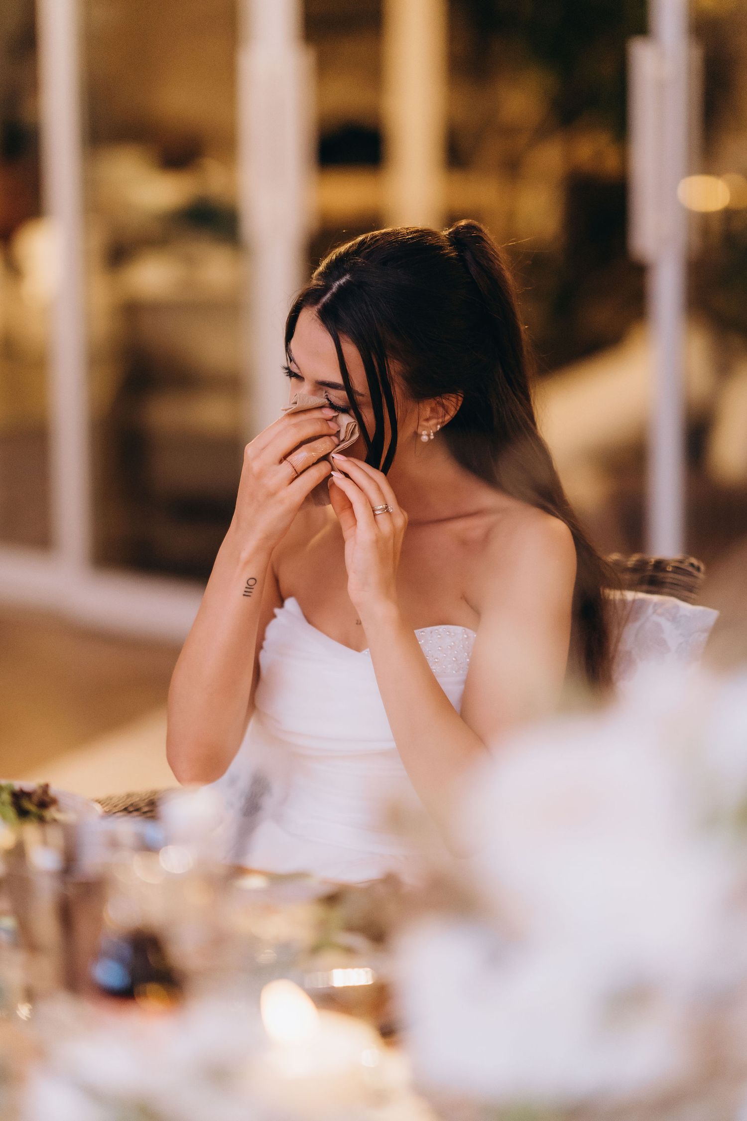 Emotional bride in white strapless gown wiping tears at wedding reception table with floral centerpieces.
