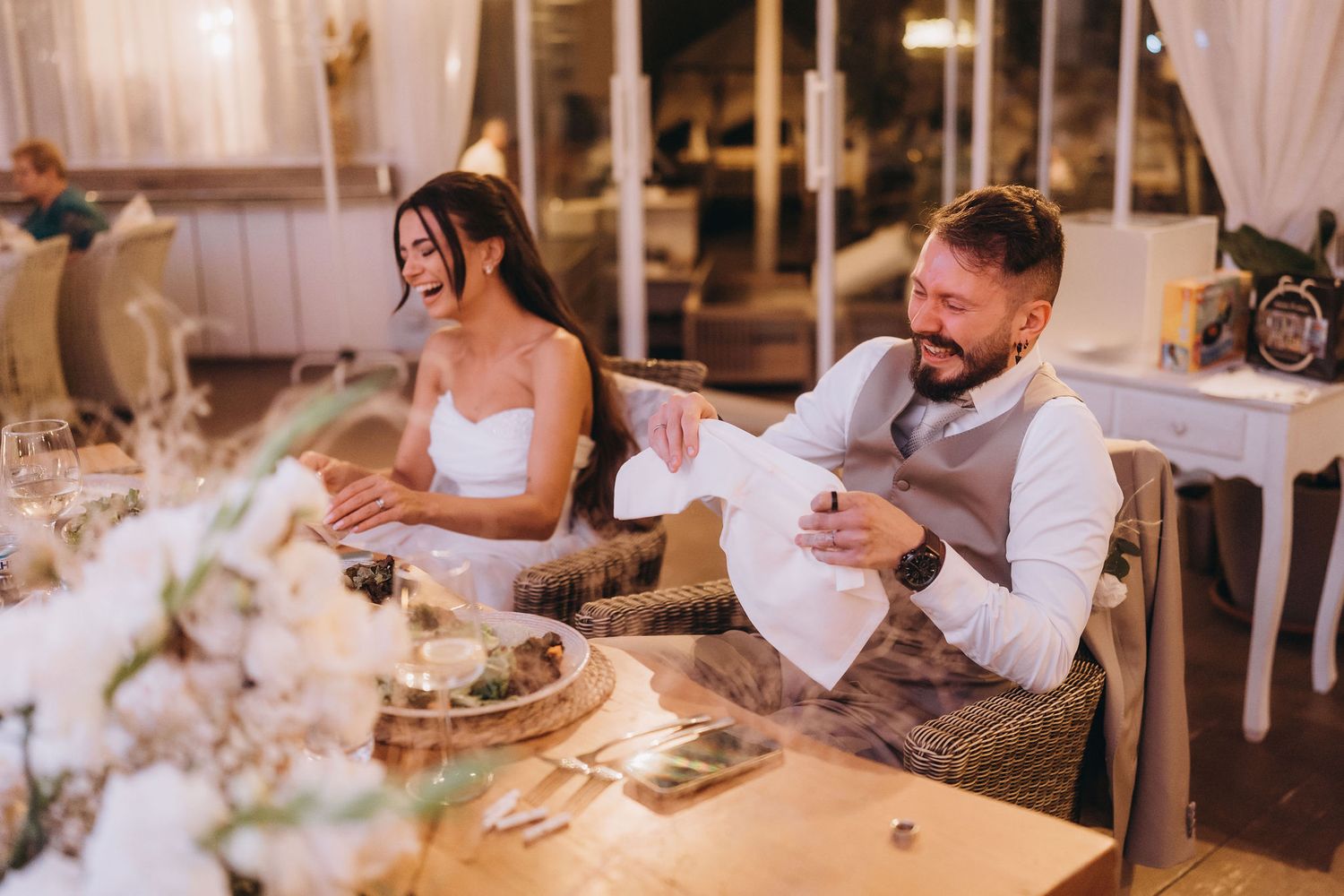 Bride and groom laughing joyfully at wedding reception table with floral centerpieces and elegant decor.