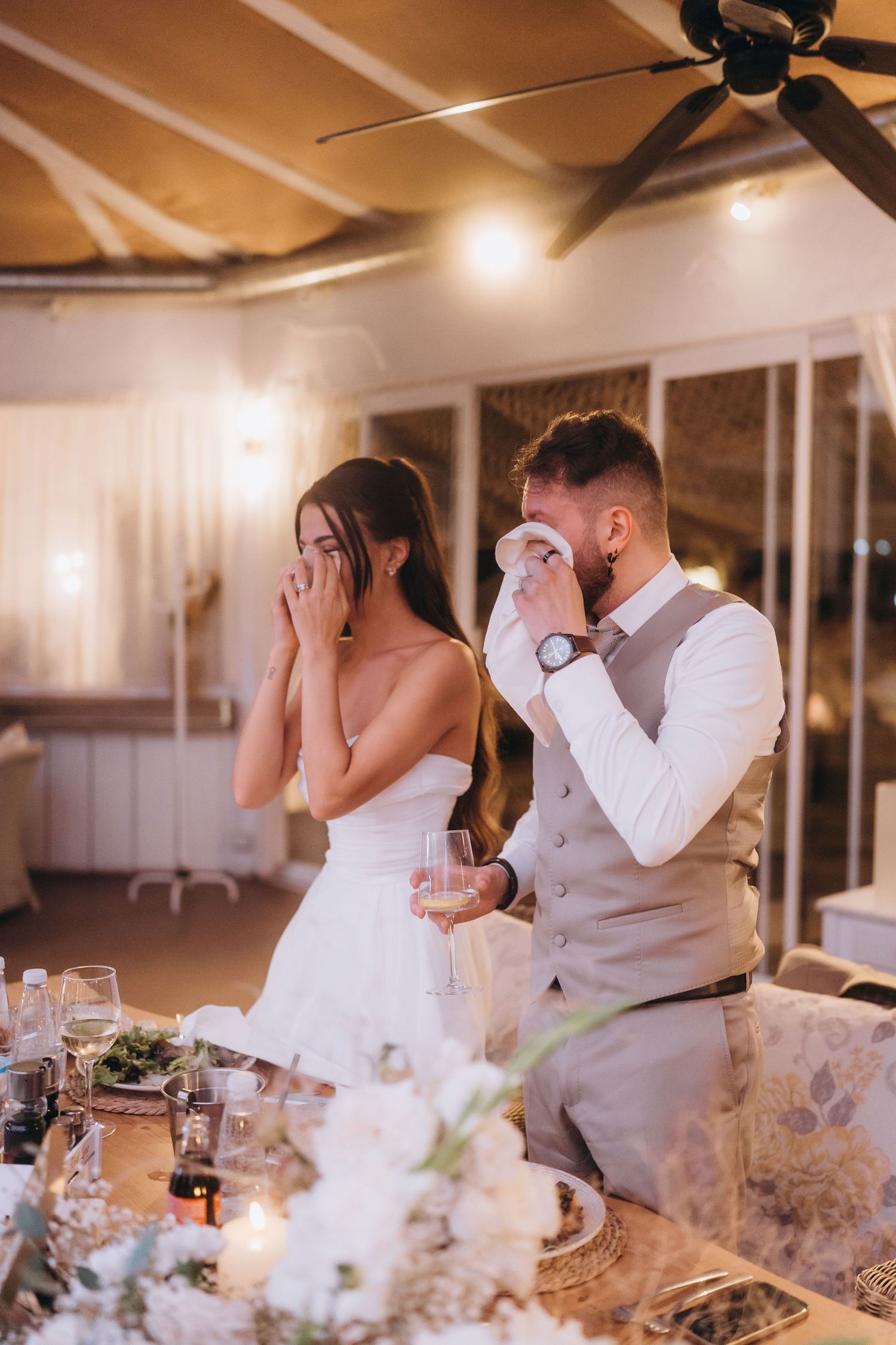 Emotional bride and groom crying during wedding reception speeches at elegantly decorated venue with warm lighting.