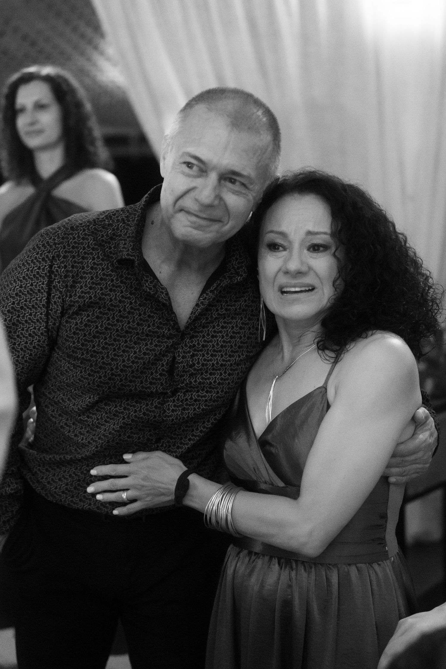 Black and white photo of a smiling couple embracing at a social event, woman in dress, man in patterned shirt.